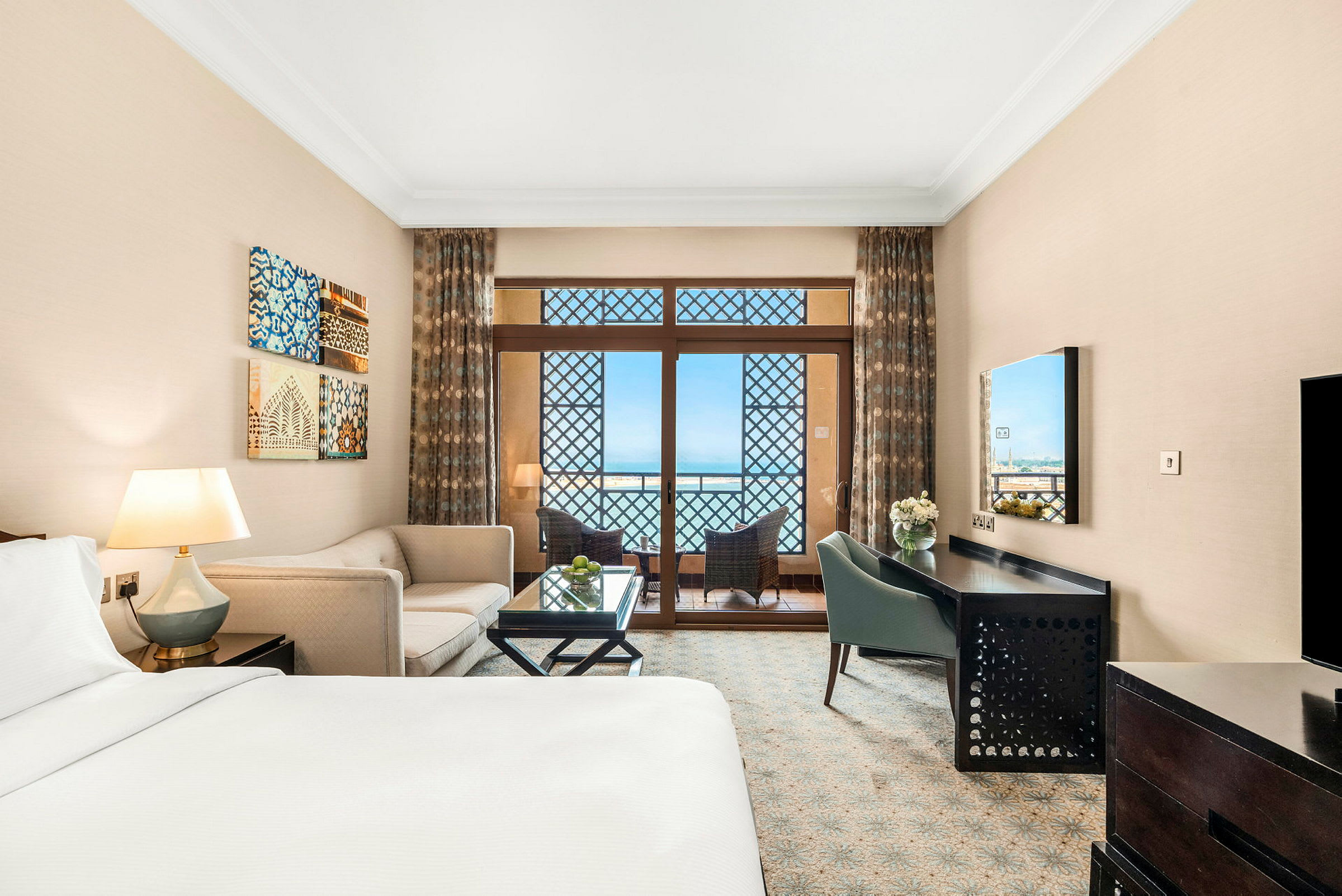 Ett hotellrum med en säng, soffa och skrivbord. Ett stort fönster leder till en balkong med utsikt över havet. (AI-genererad text)