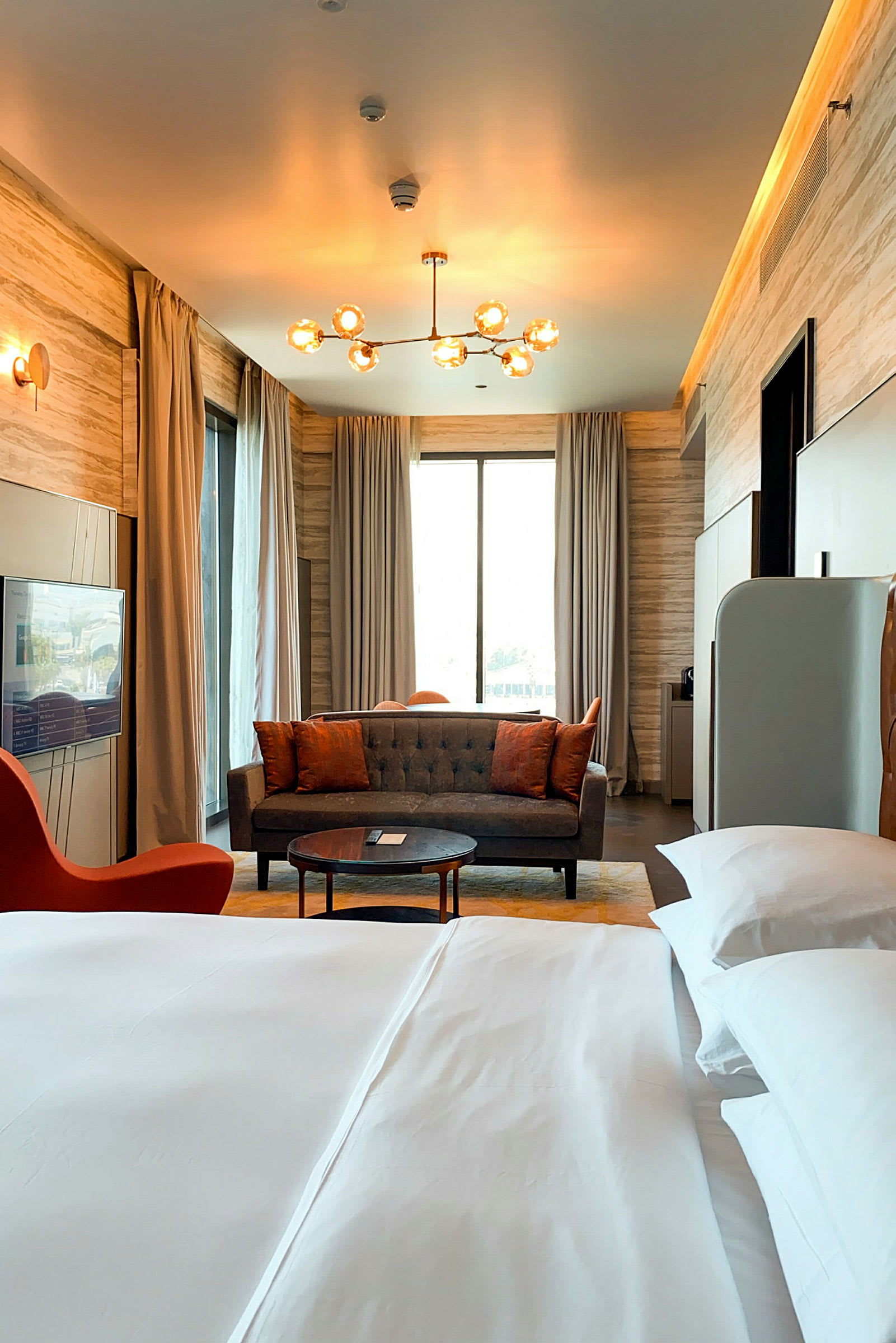 Ett elegant möblerat hotellrum med en stor säng i förgrunden och en soffgrupp framför ett stort fönster med gardiner. En modern lampa hänger i taket. (AI-genererad text)