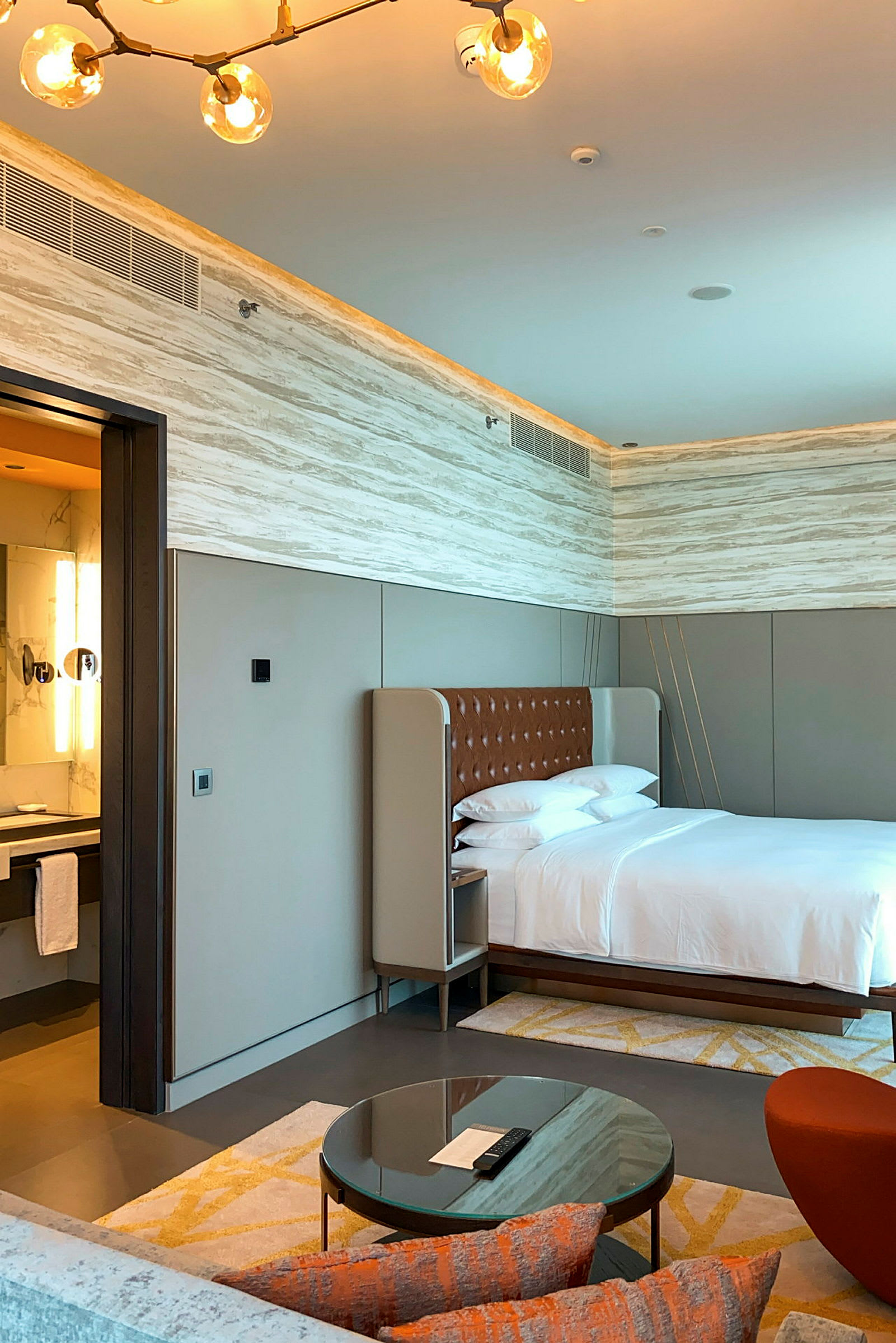 Sovrum i hotell med en stor säng, ett angränsande badrum, en sittgrupp med en glasbord och en modern belysning i taket. (AI-genererad text)