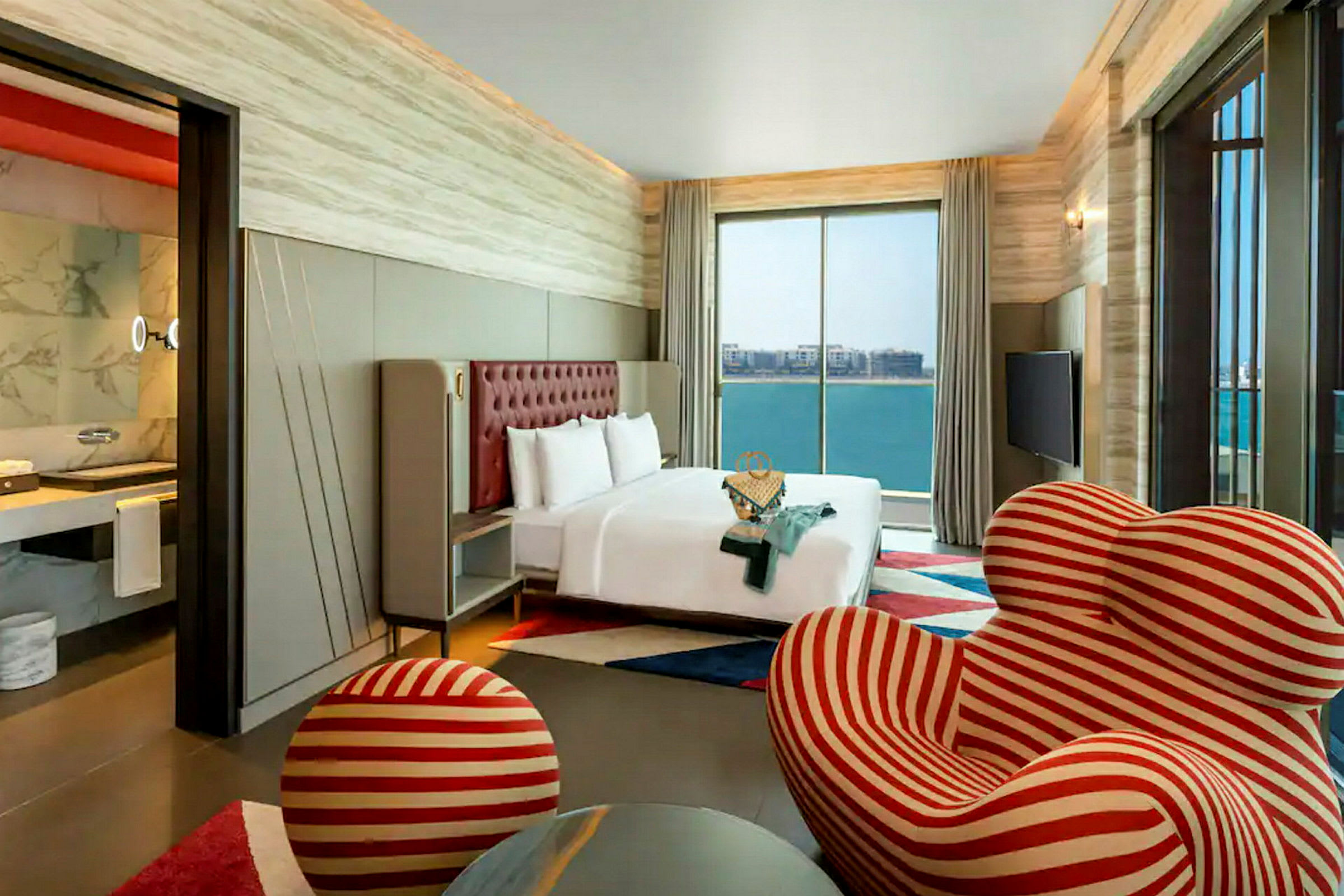 Ett modernt hotellrum med en stor säng framför ett stort fönster som visar utsikt över havet. Rummet har också ett unikt randigt rött och vitt fåtölj med en matchande fotpall. (AI-genererad text)