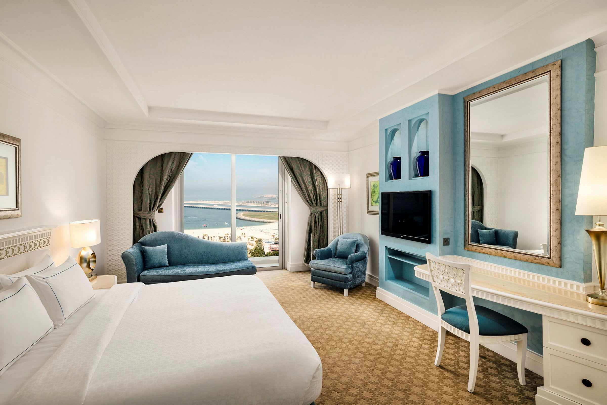 Hotellrum med säng, soffor, skrivbord och spegel. Utsikt mot havet genom fönstret. (AI-genererad text)