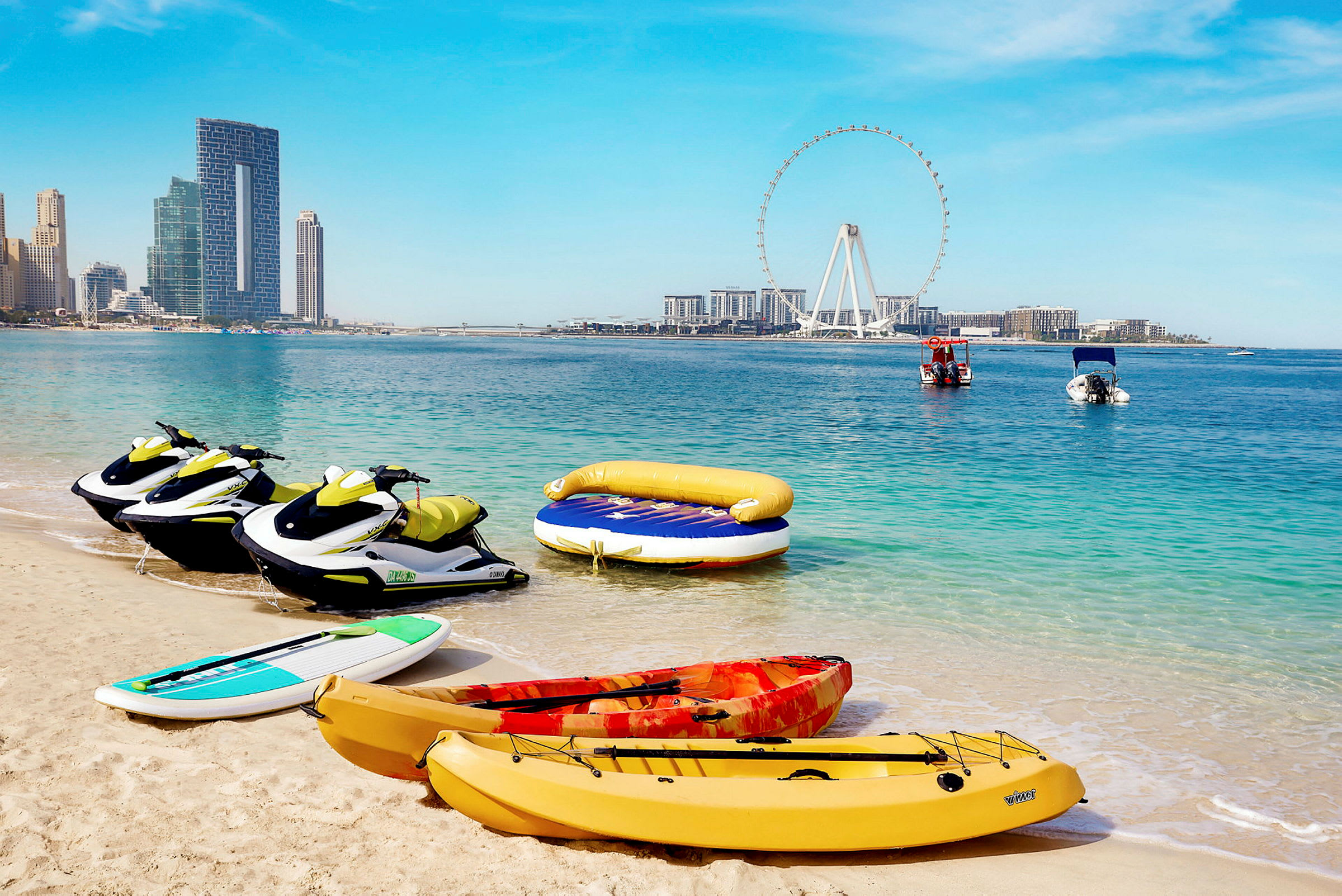 Jetski, kajaker och en paddleboard vid stranden med havet och stadssiluetten i bakgrunden. (AI-genererad text)