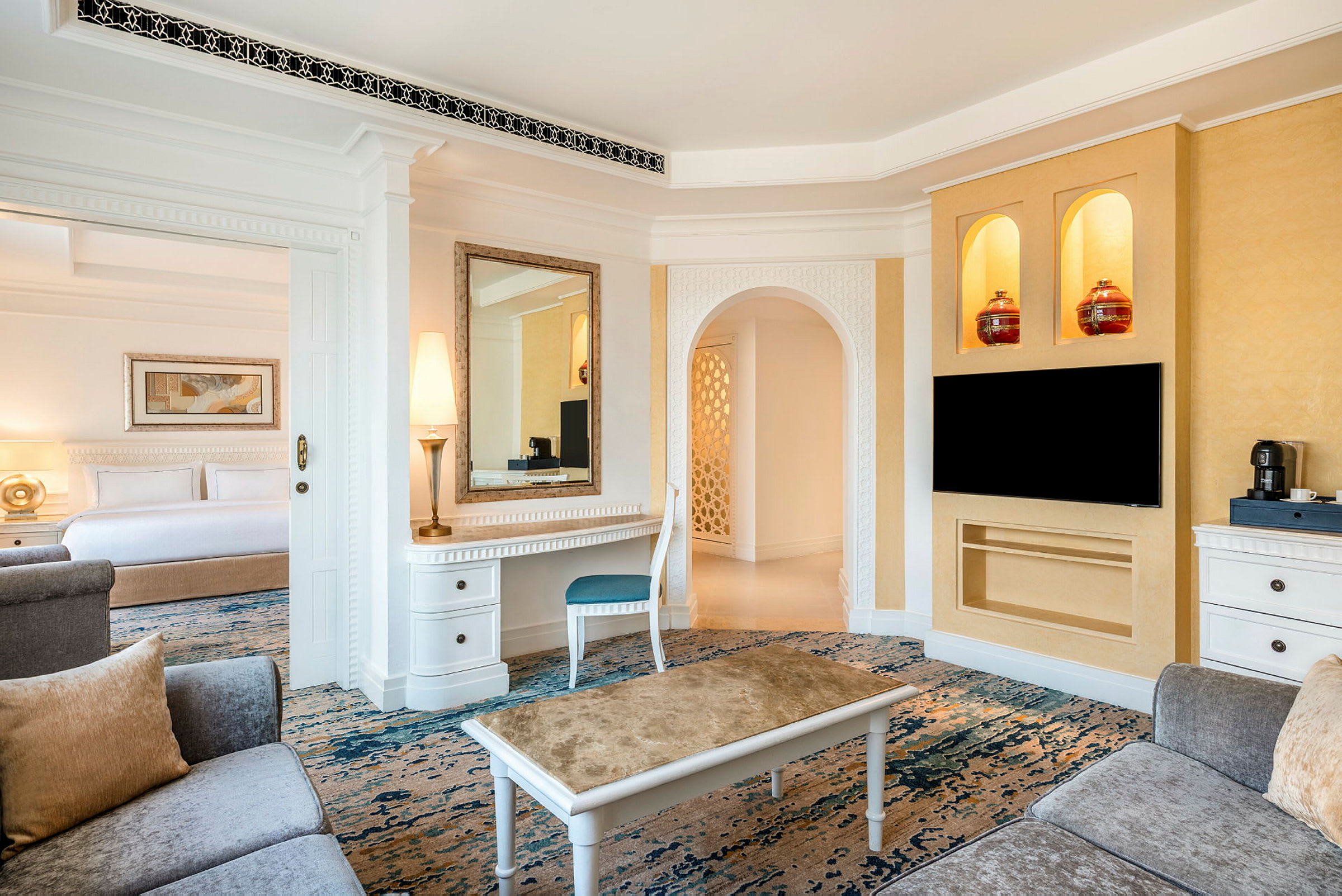 Ett elegant hotellrum med vardagsrumsmöbler, ett skrivbord med spegel och lampa, samt en öppen anslutning till sovrummet med en stor säng. TV på väggen och soffmöbler framför. (AI-genererad text)