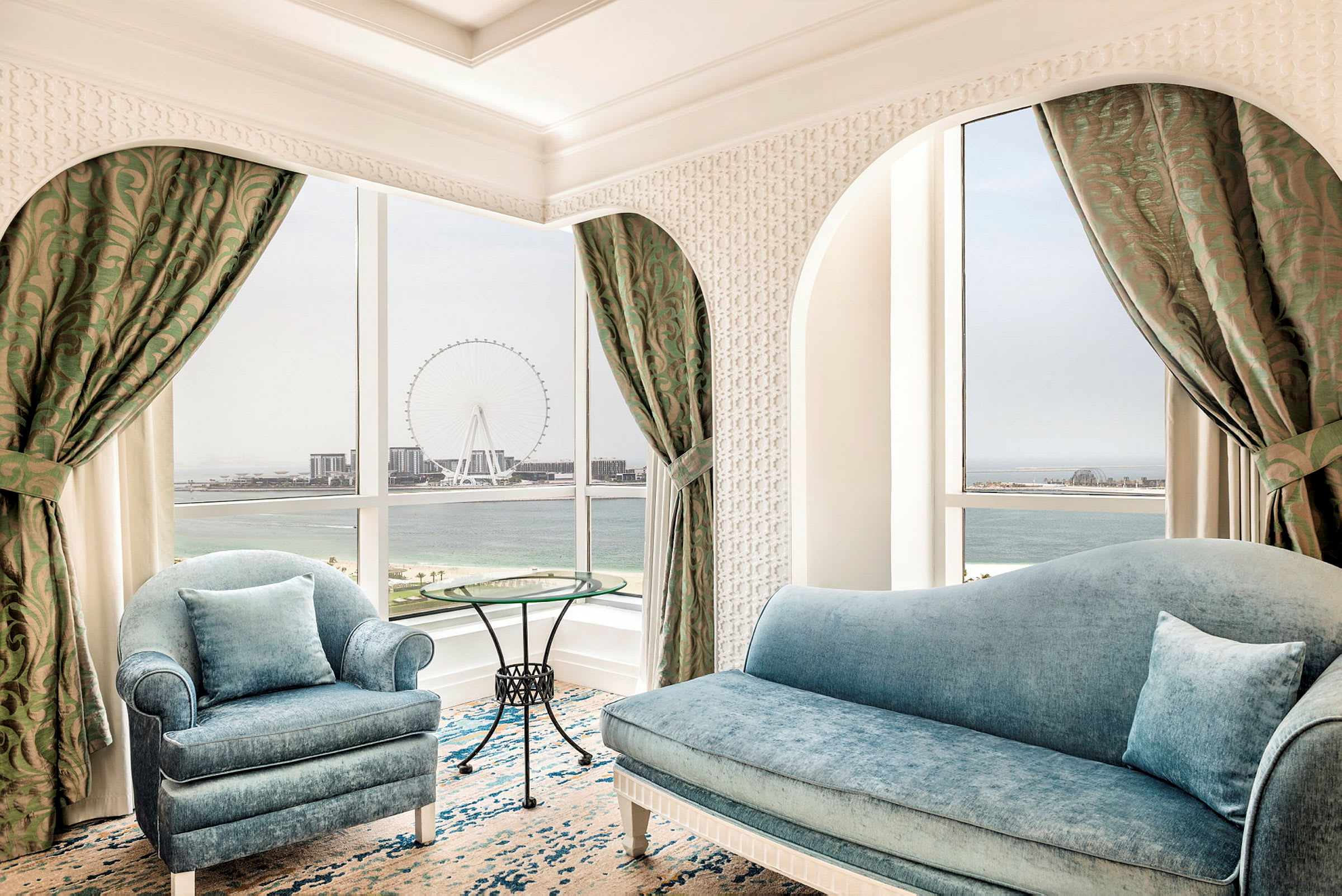 Ett elegant rum med blå soffor och ett runt glasbord. Fönstret har utsikt över havet och ett stort pariserhjul. (AI-genererad text)