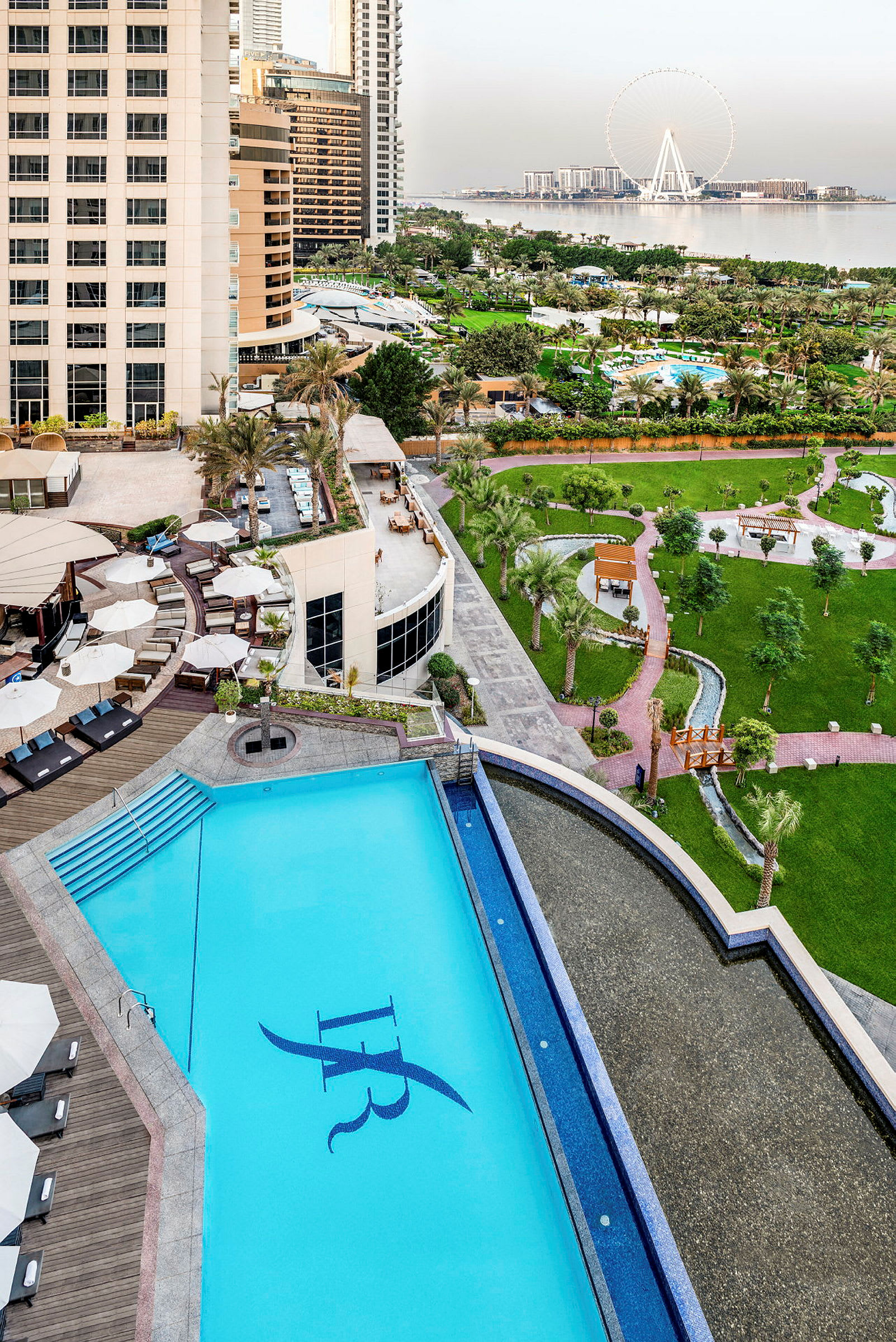Ett hotellområde med pool, grönområden och utsikt över havet och ett stort pariserhjul. (AI-genererad text)