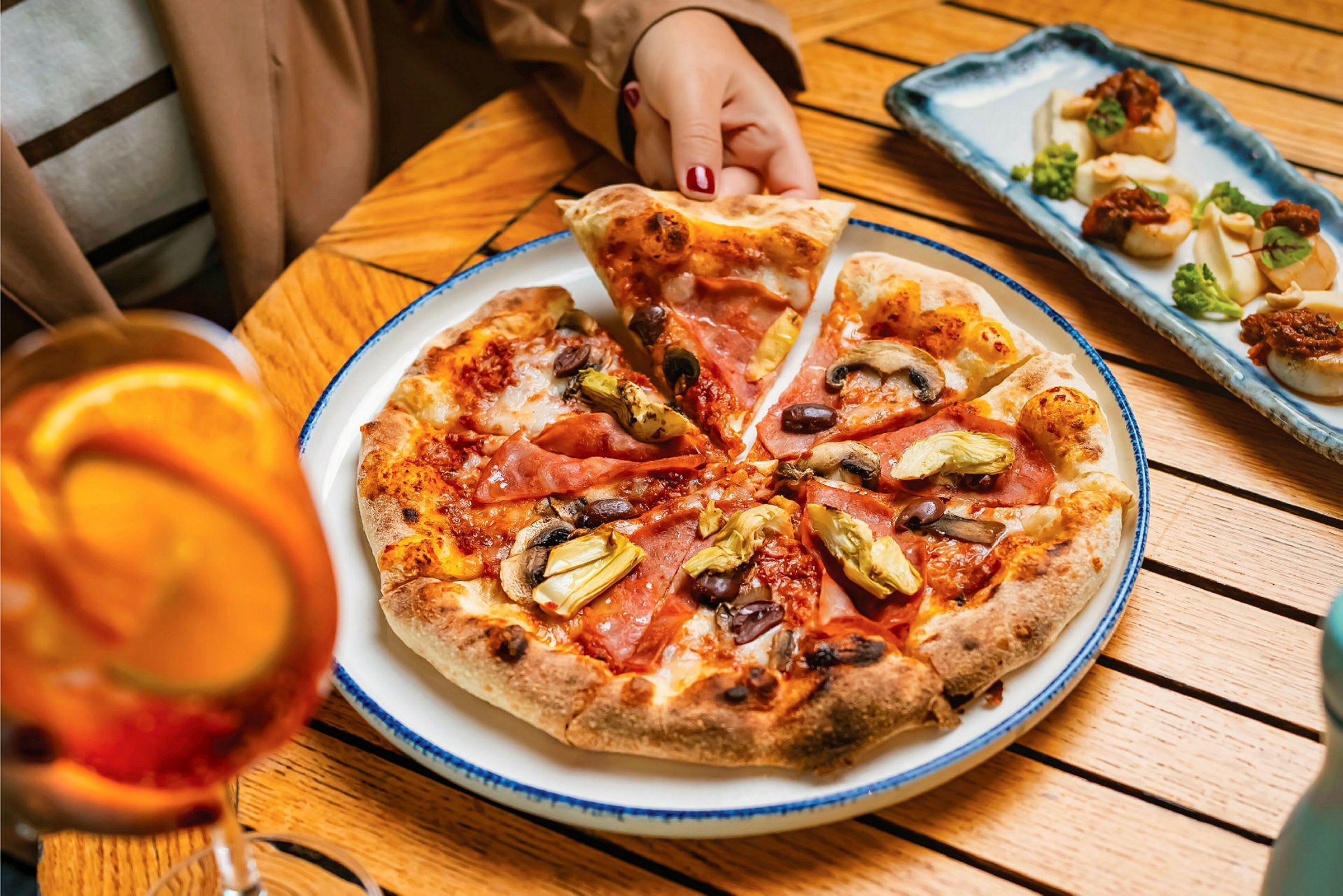 En tallrik med pizza toppad med skinka, oliver, svamp och kronärtskockor på ett bord. En person tar en pizzabit. Bakom pizza finns en kurv med små förrätter och bredvid ett glas med orange dryck. (AI-genererad text)