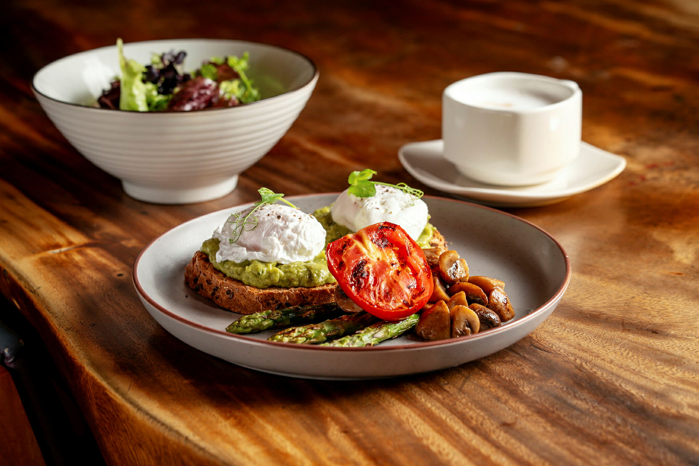 En tallrik med toast, mosad avocado, pocherade ägg, grillad tomat, sparris och svamp. Bakom syns en salladsskål och en kopp på ett träbord. (AI-genererad text)