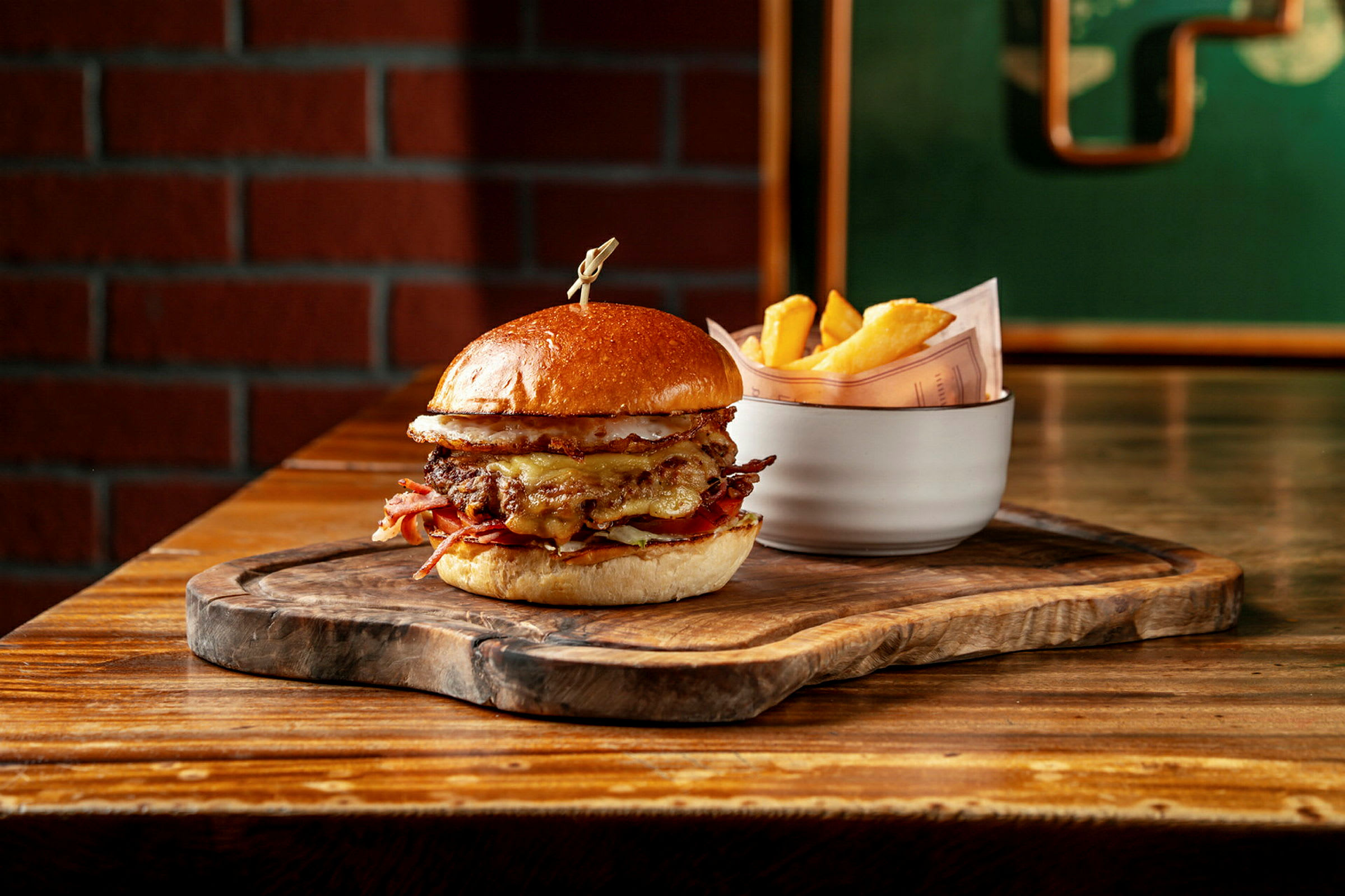 En hamburgare med bröd, kött och ost serverad på ett träunderlägg. Bredvid står en vit skål med pommes frites. (AI-genererad text)