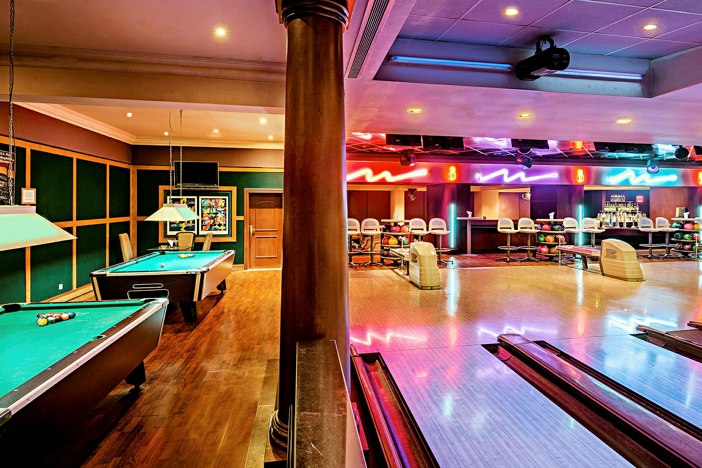 Ett rum med biljardbord på vänster sida, en bowlingbana till höger och ett område med barstolar och flaskor i bakgrunden. (AI-genererad text)