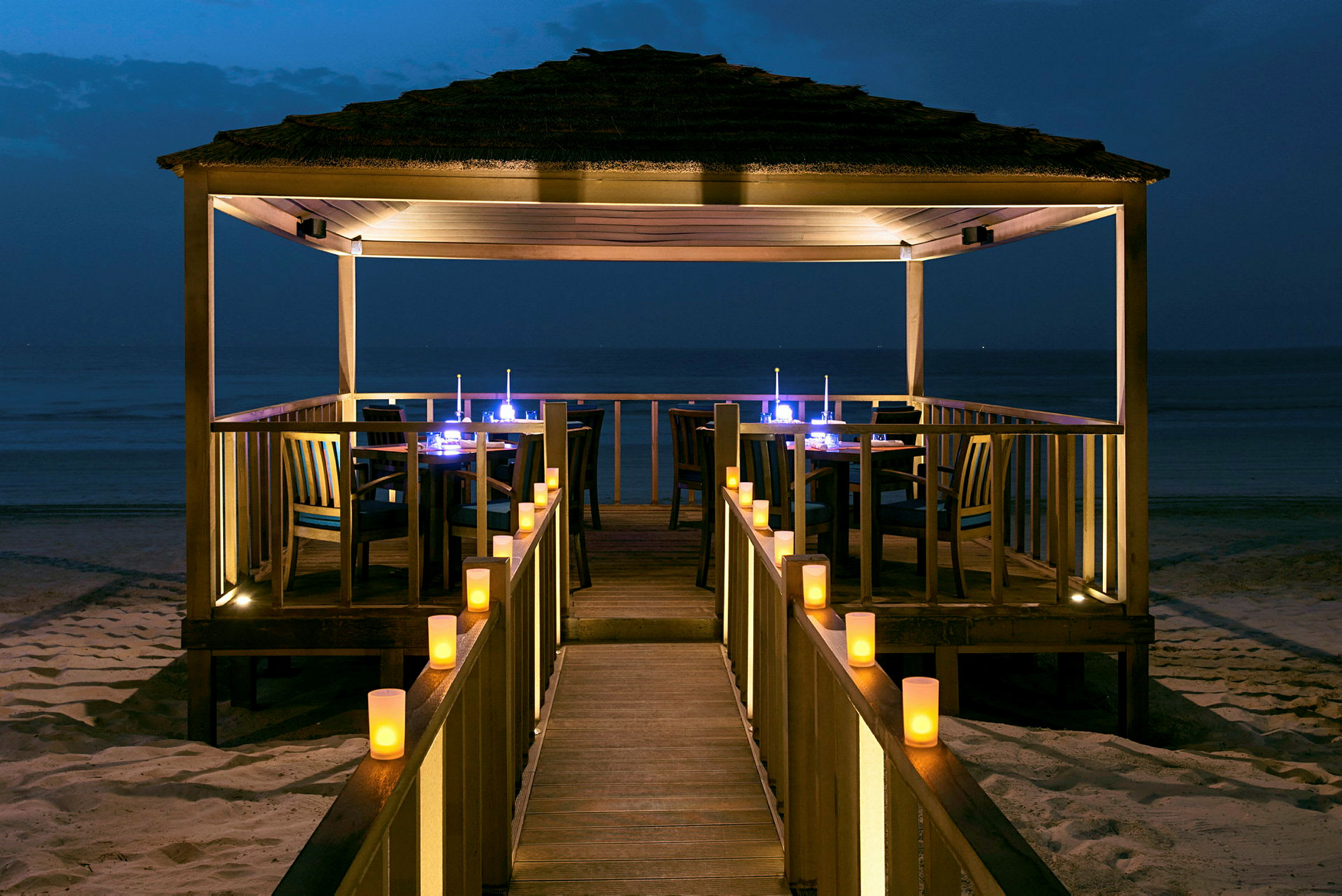 Ett lyst träpaviljong på en sandstrand med ljusdekorationer, med havet som bakgrund i skymningen. (AI-genererad text)