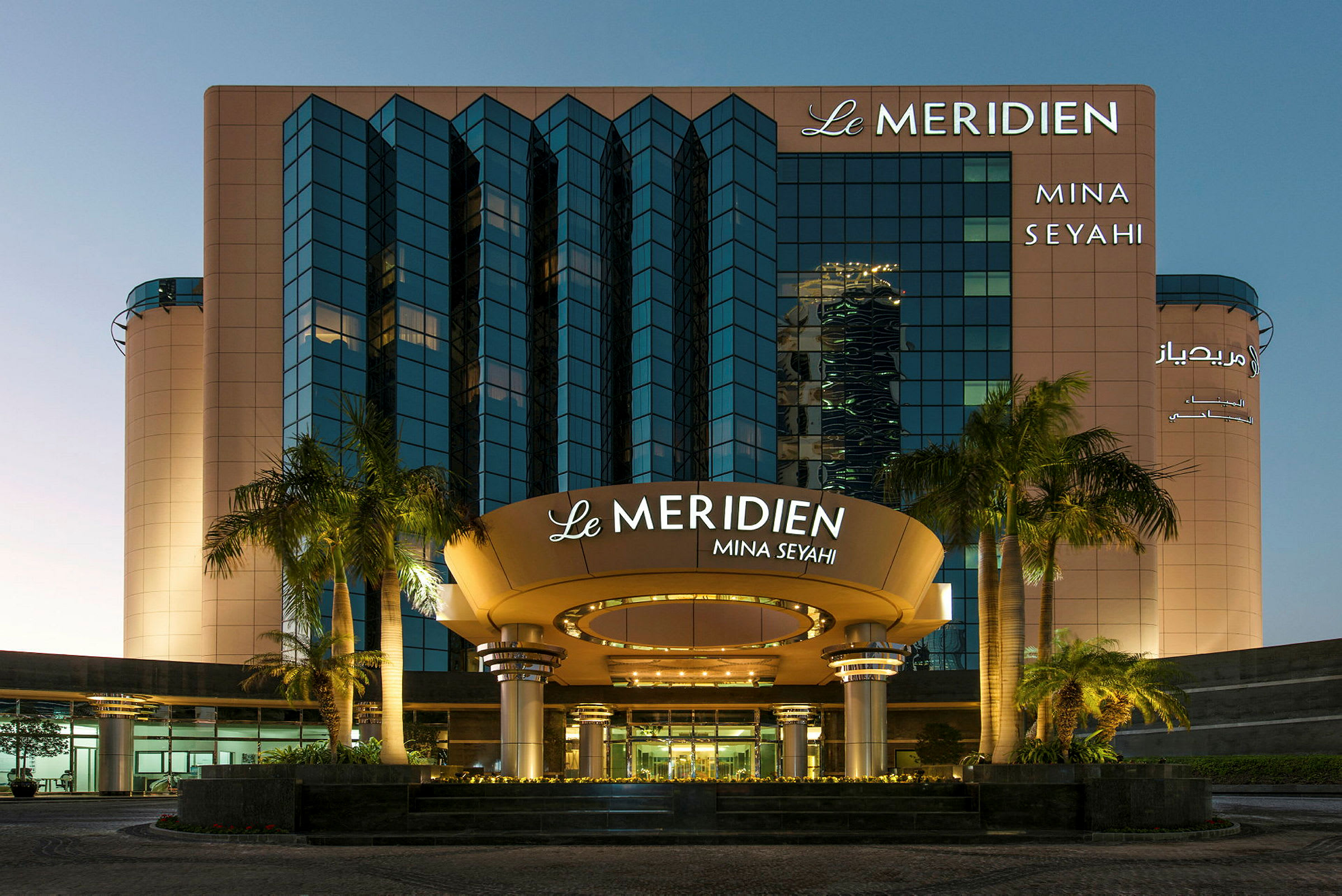 Fasaden av Le Meridien Mina Seyahi hotell med palmer i förgrunden. (AI-genererad text)