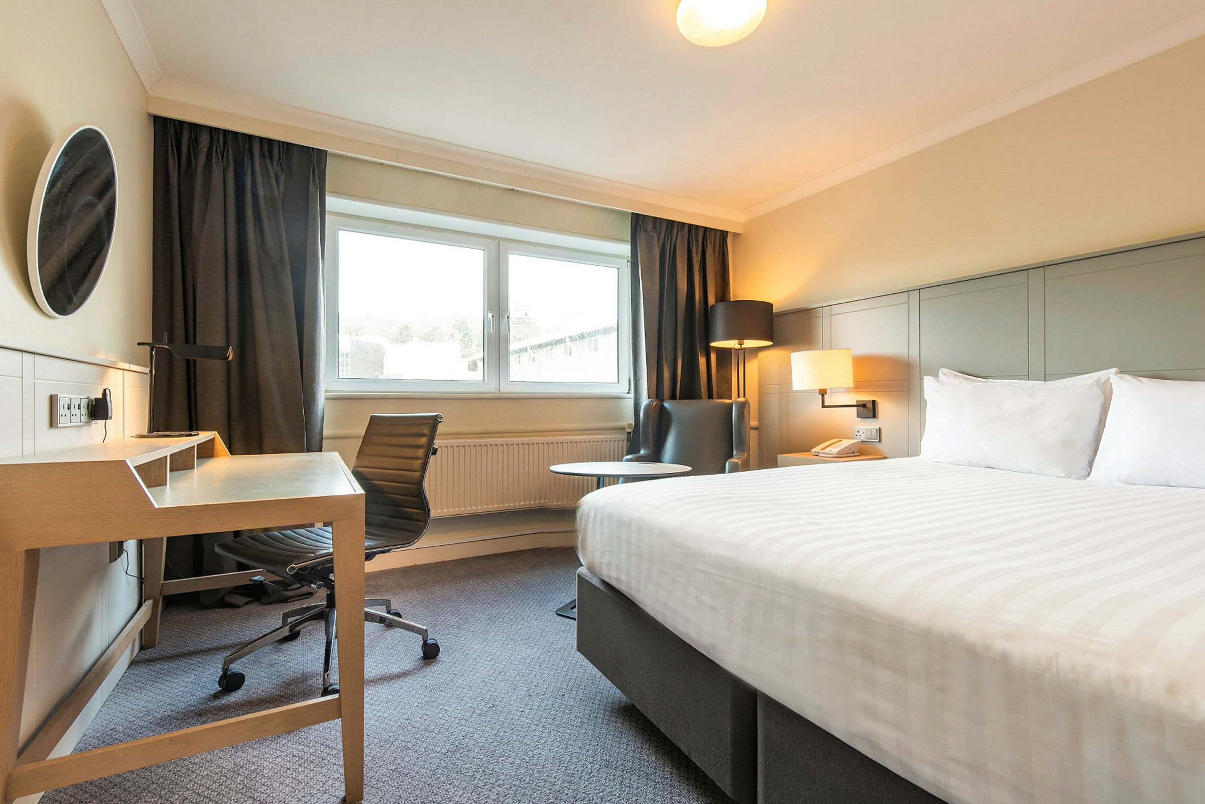 Bild på ett hotellrum med en säng, skrivbord och stol, och en fåtölj bredvid ett fönster. (AI-genererad text)