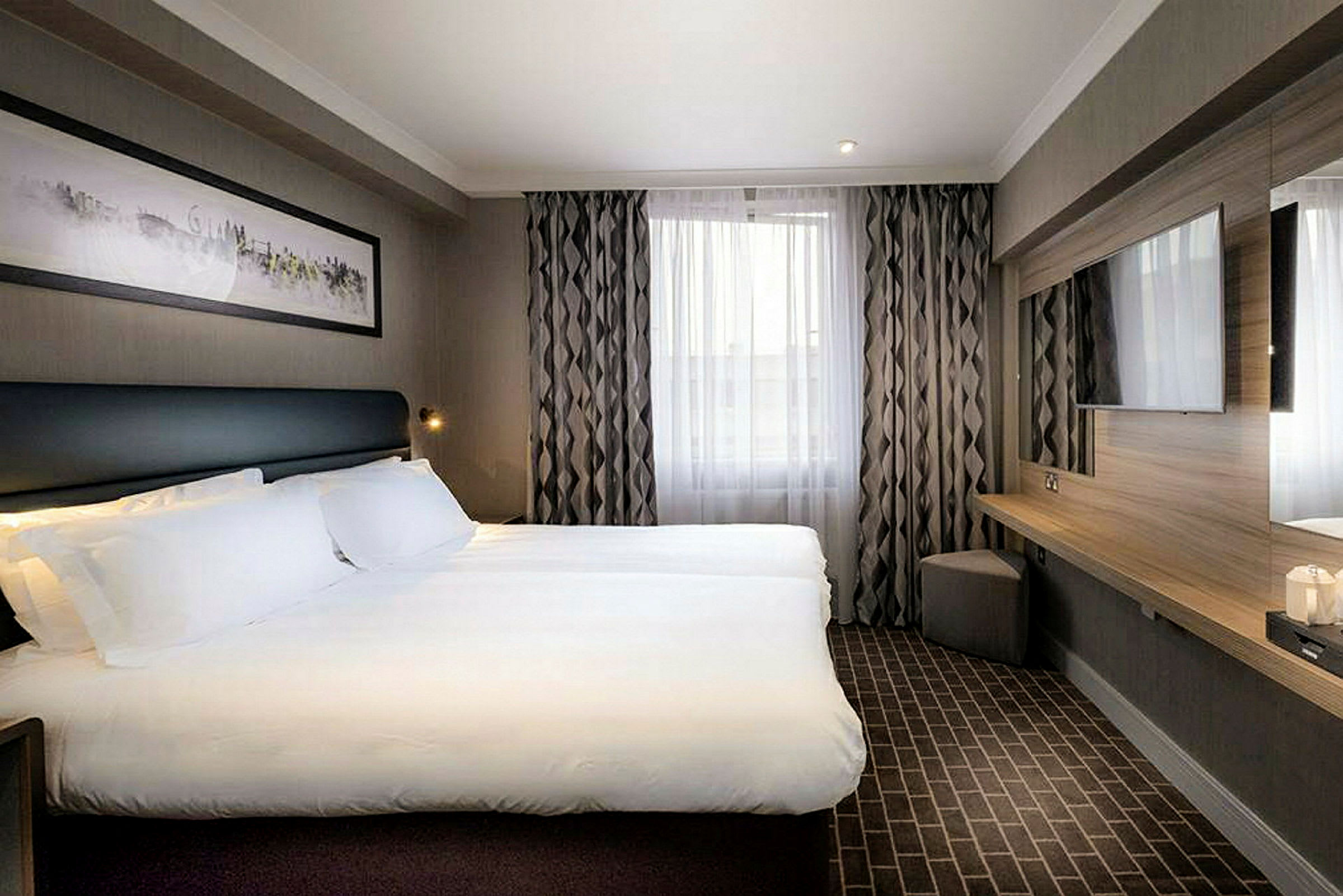 Modernt hotellrum med en dubbelsäng, väggmonterad TV och fönster med gardiner. (AI-genererad text)