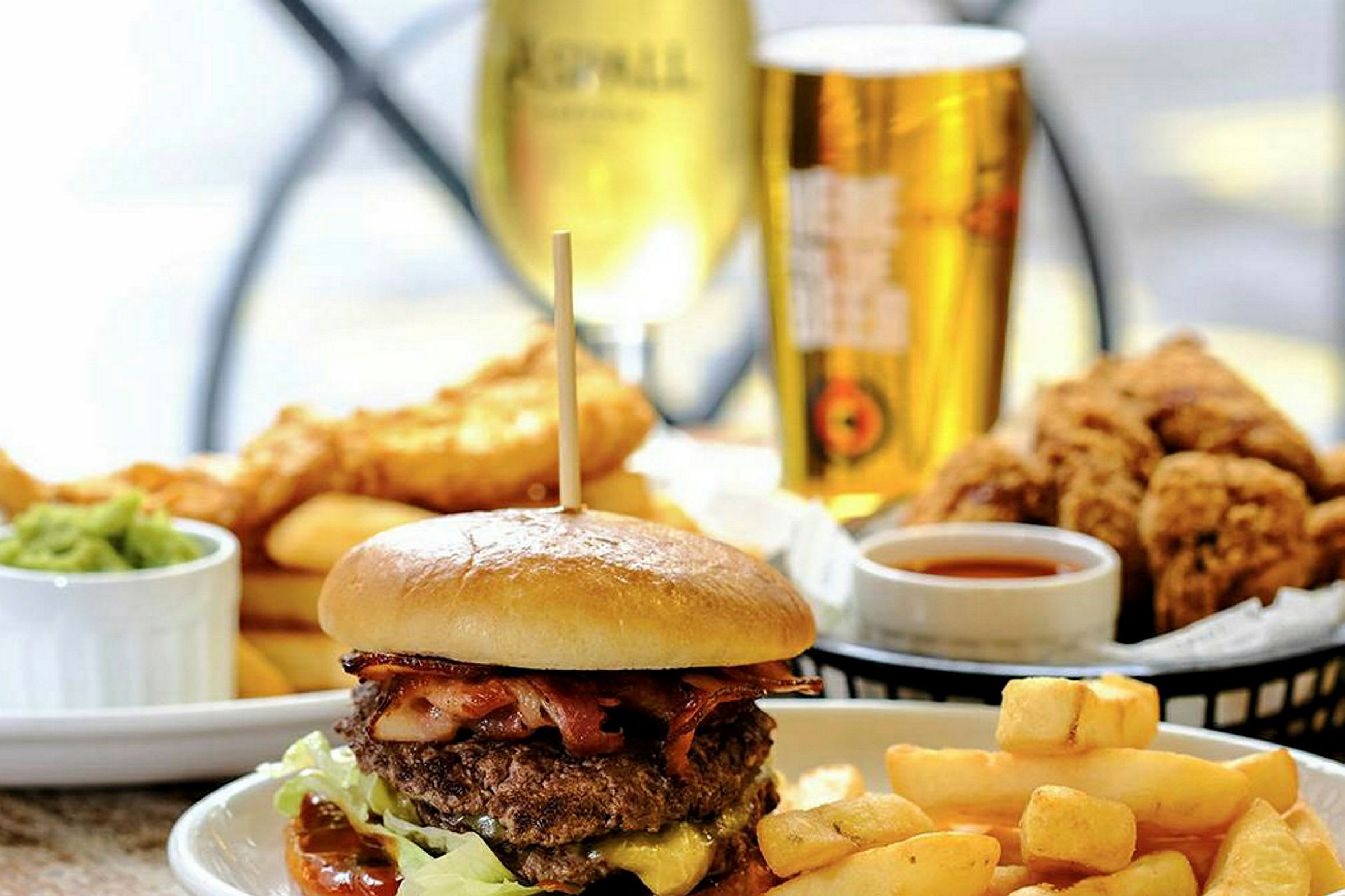 En köttburgare med pommes frites och tillbehör, dryck och friterad mat i bakgrunden. (AI-genererad text)