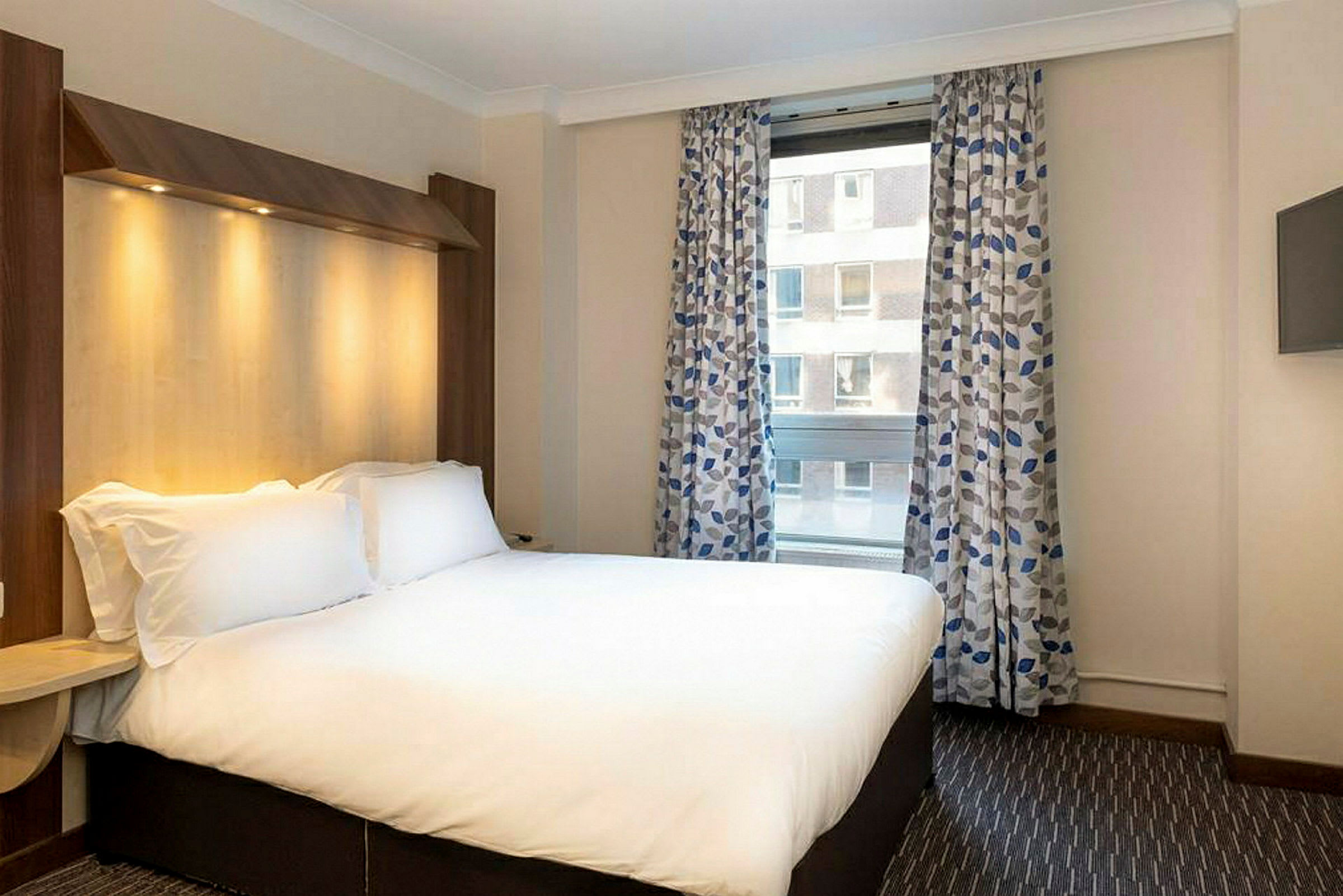 Ett hotellrum med en dubbelsäng, ett fönster med mönstrade gardiner och en väggmonterad TV. (AI-genererad text)