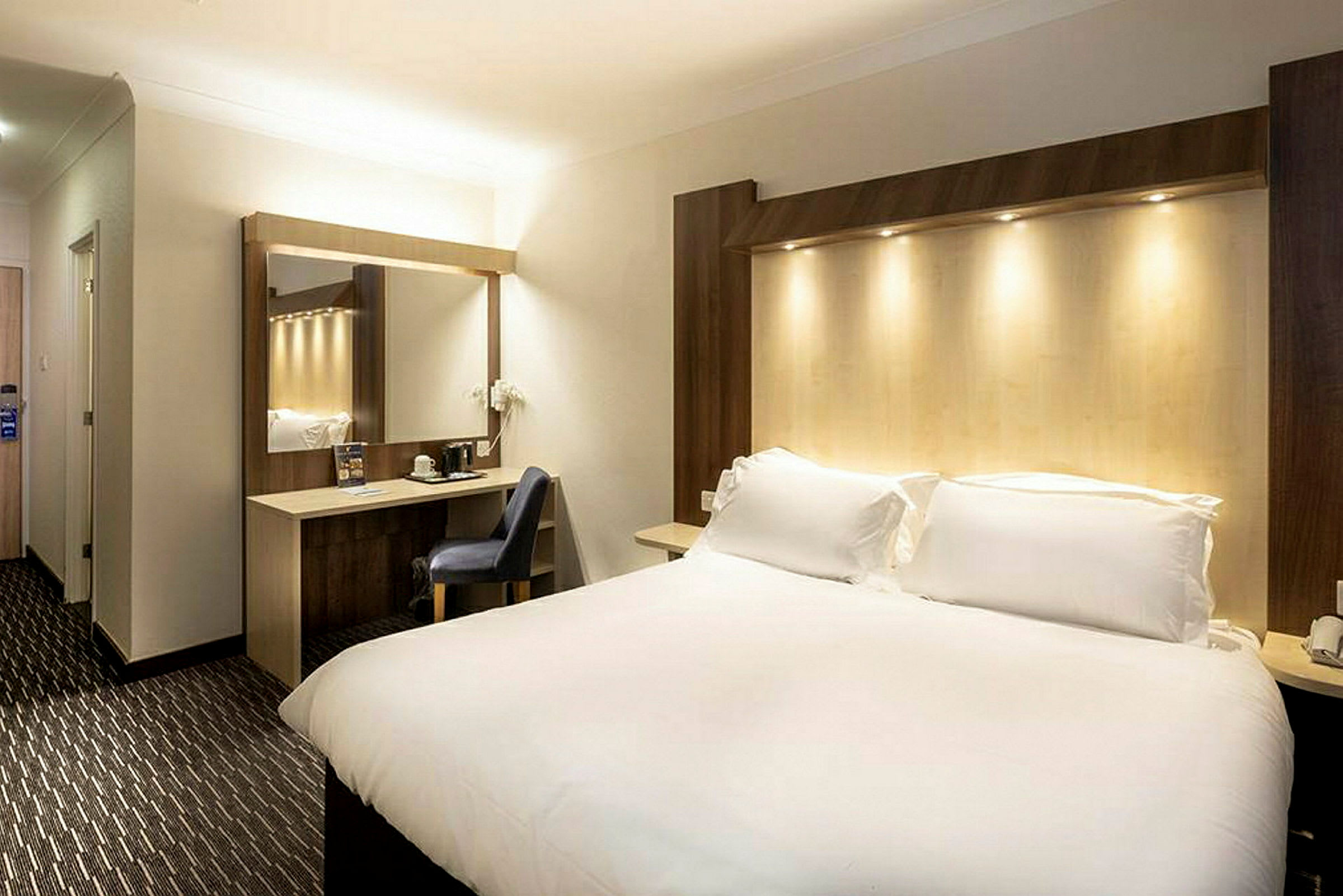 Ett hotellrum med en stor säng, en arbetsyta med stol och en spegel. (AI-genererad text)