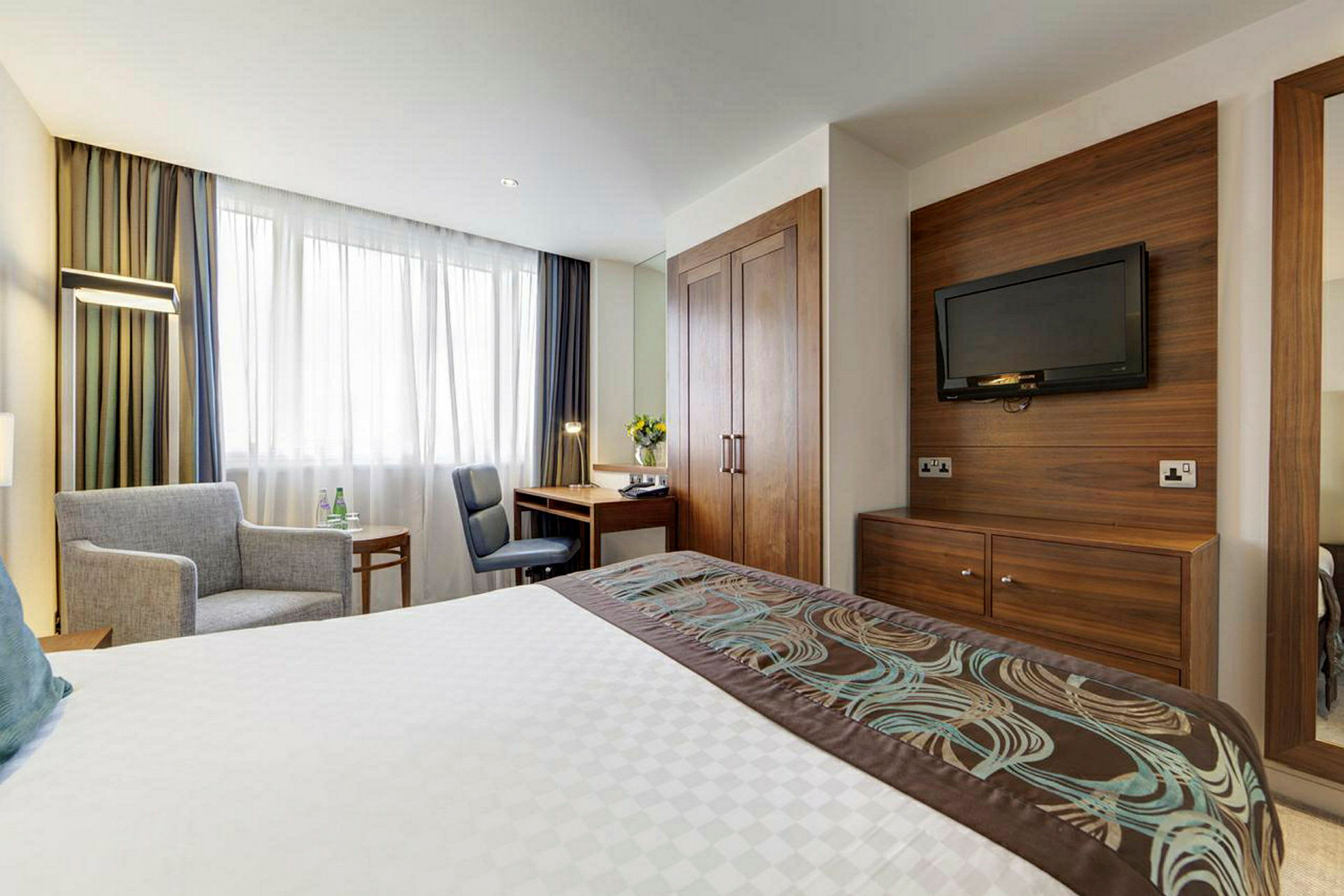 Ett hotellrum med en säng, en fåtölj, ett skrivbord och en TV på väggen. (AI-genererad text)