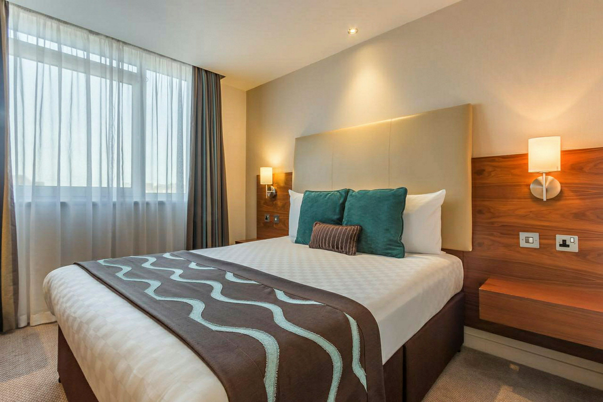 Ett hotellrum med en dubbelsäng med dekorativa kuddar och belysning på väggen. (AI-genererad text)