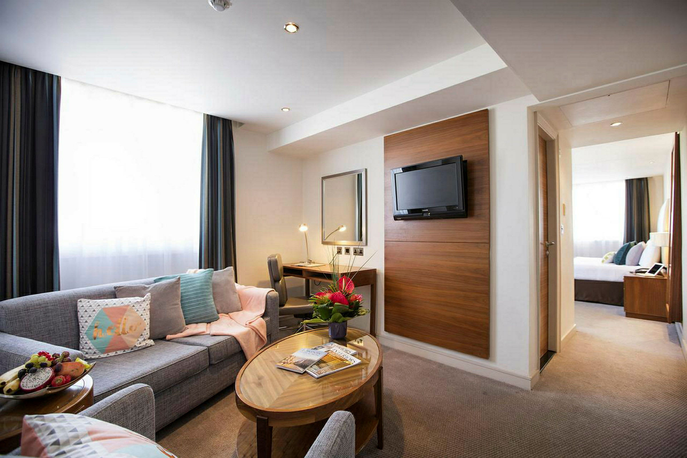 Ett hotellrum med soffgrupp, soffbord, skrivbord, väggmonterad TV och blommor. (AI-genererad text)