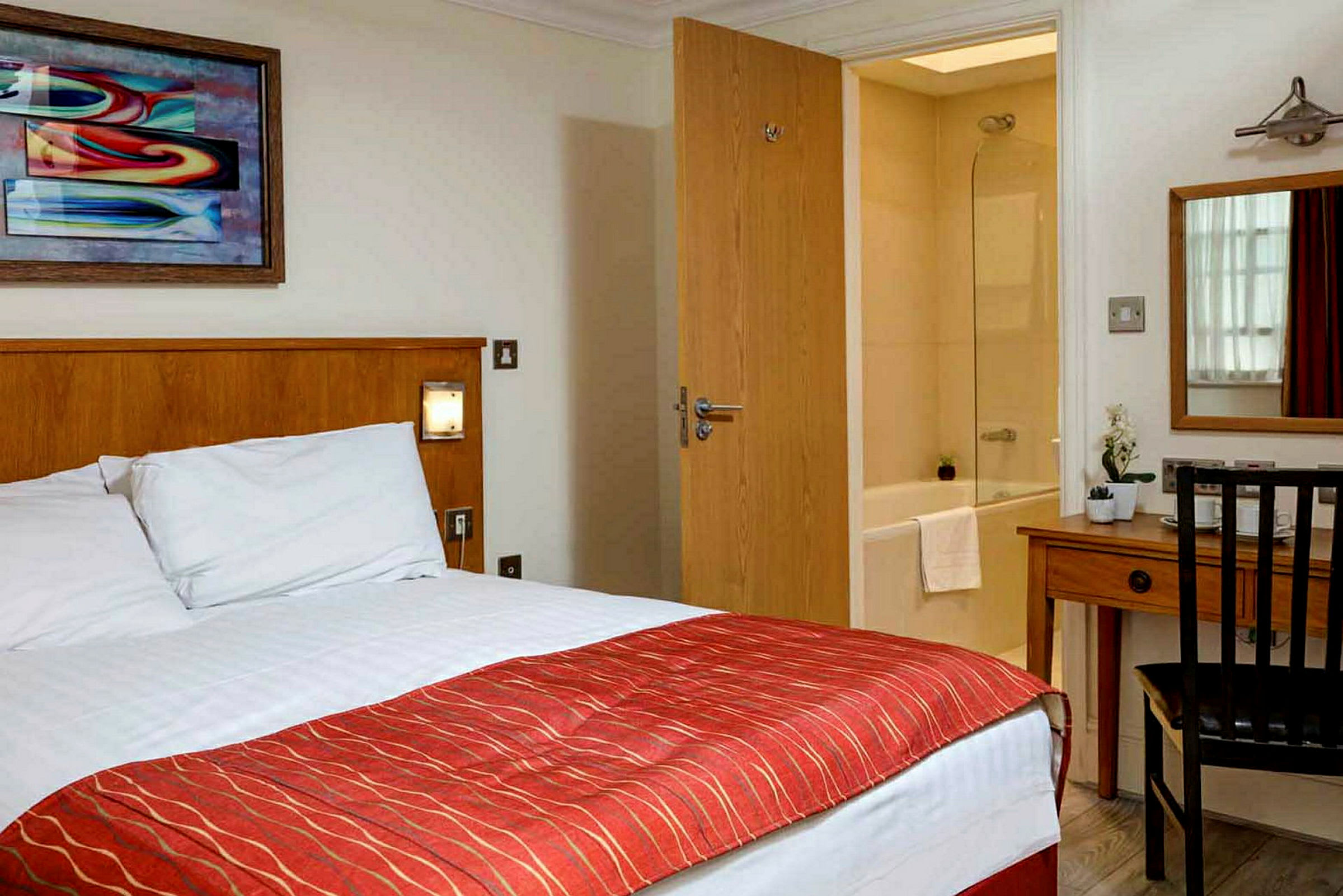 Hotellrum med säng, skrivbord, spegel och vy mot ett badrum. (AI-genererad text)