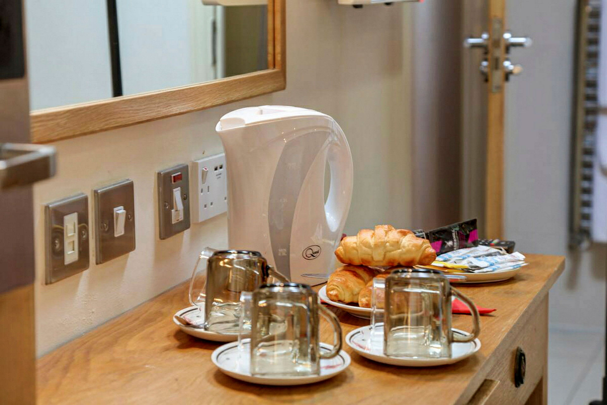 Vattenkokare, kaffekoppar med fat, croissanter och kaffepåsar på en träbänk. (AI-genererad text)