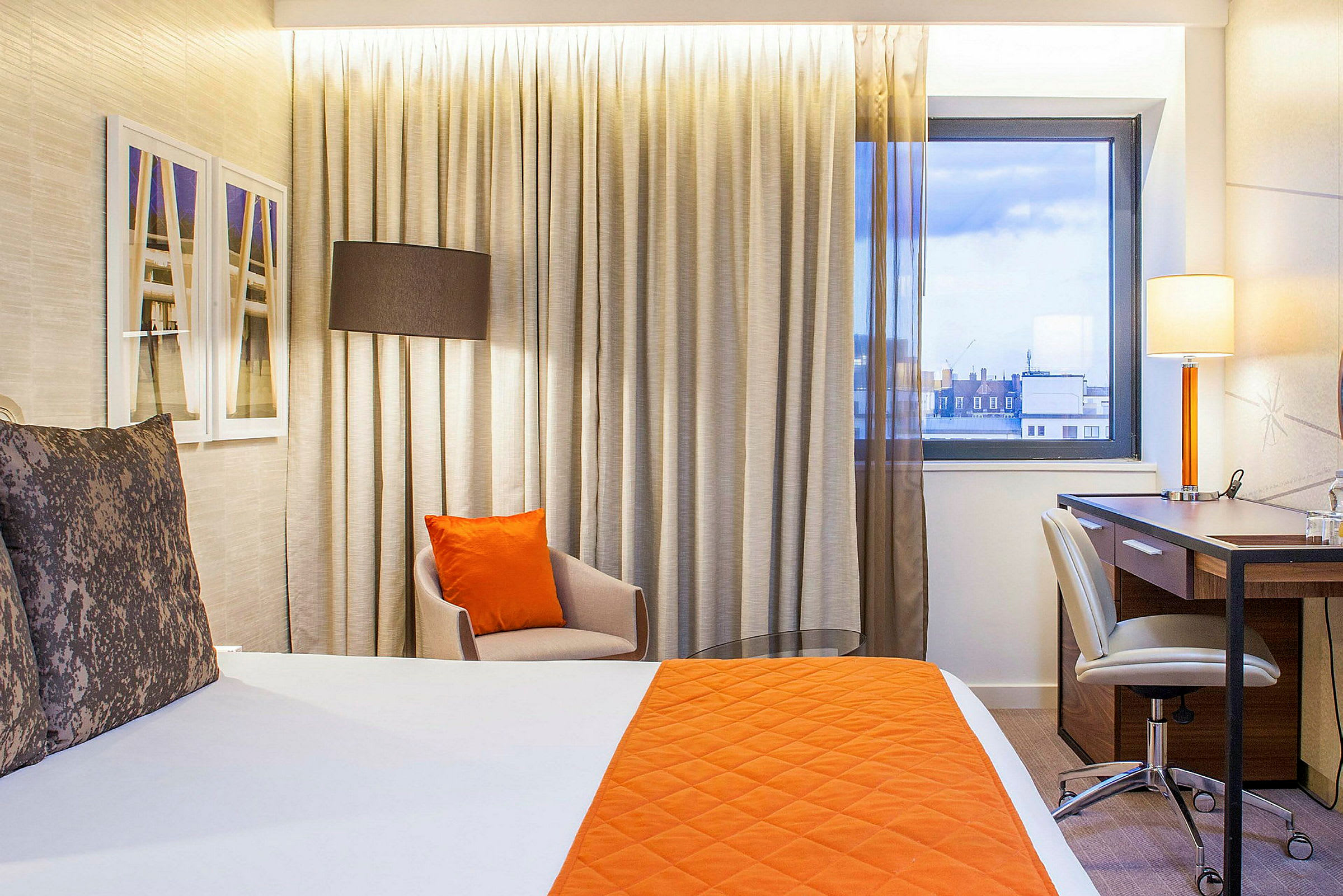 Ett hotellrum med ett sängöverkast och kudde i orange färg, en stol, och ett skrivbord nära fönstret. (AI-genererad text)