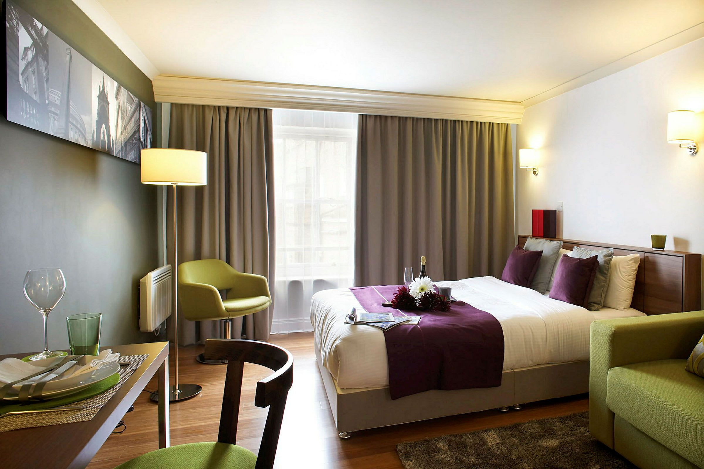 Ett hotellrum med en dubbelsäng klädd i lila och vita sängkläder, en grön stol och ett matbord med porslin. (AI-genererad text)
