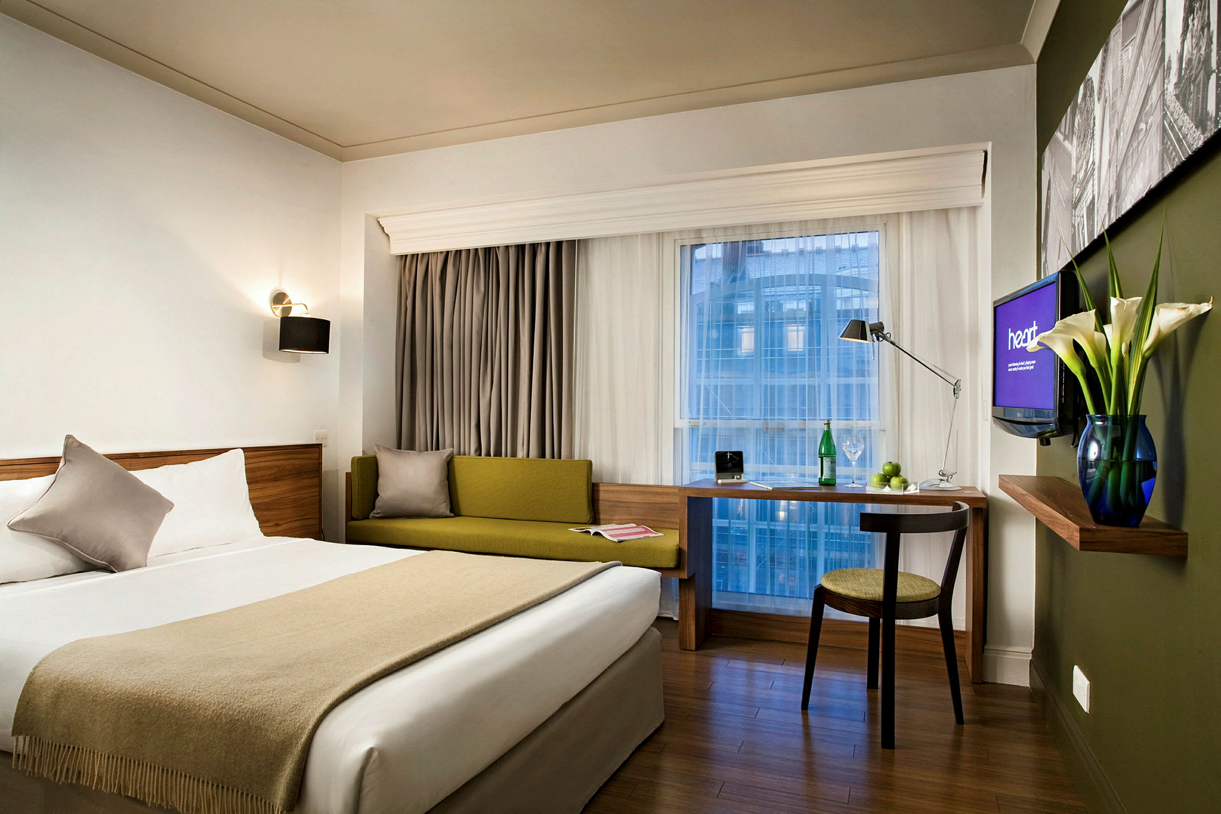 Ett modernt hotellrum med en dubbelsäng, grön soffa, skrivbord och en stor fönstervy. (AI-genererad text)