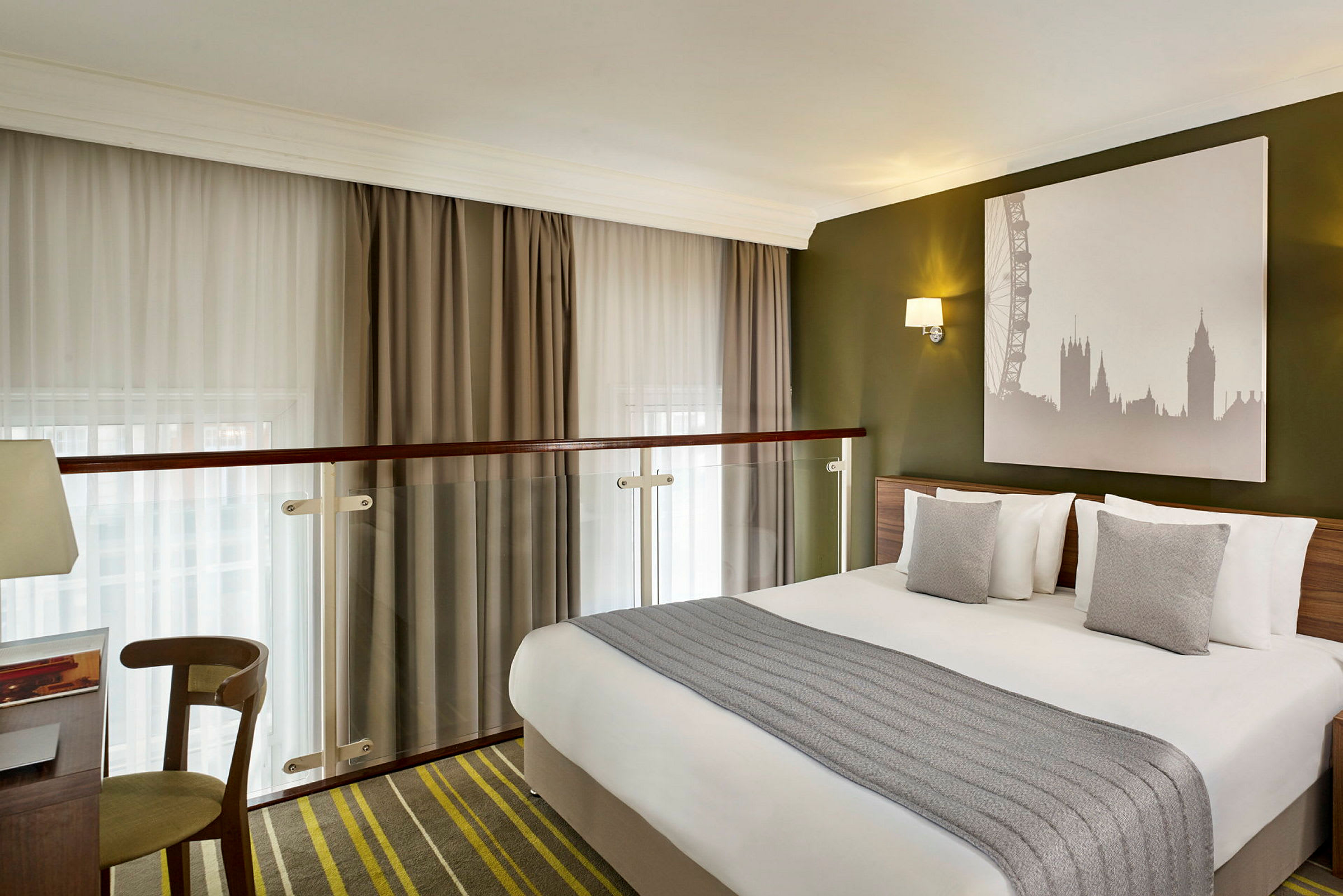 Ett hotellrum med en dubbelsäng, gråa kuddar, och en bild av Londons skyline på väggen. (AI-genererad text)