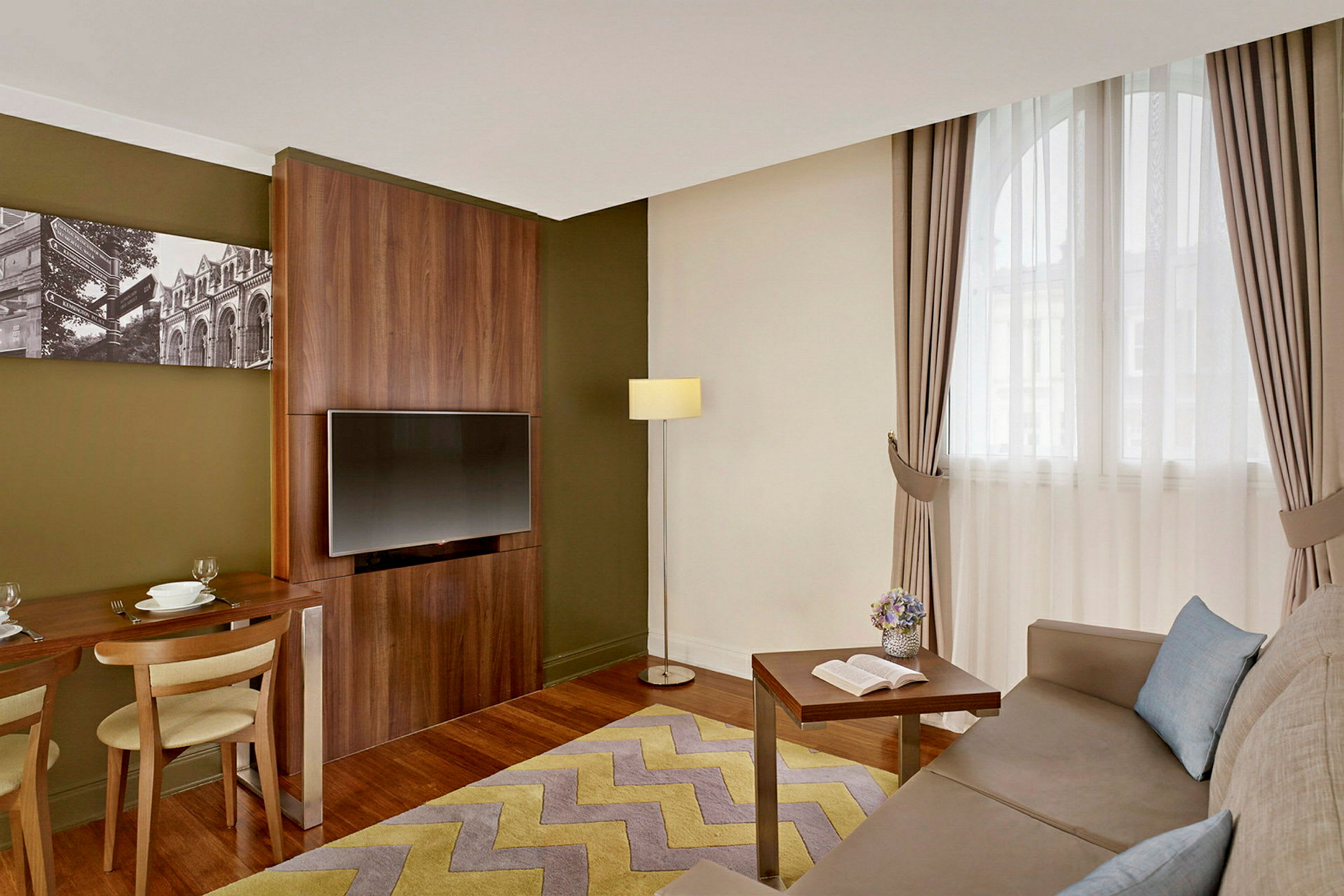 Interiör av ett hotellrum med en soffa, ett soffbord, en TV på väggen och ett matbord med två stolar. (AI-genererad text)