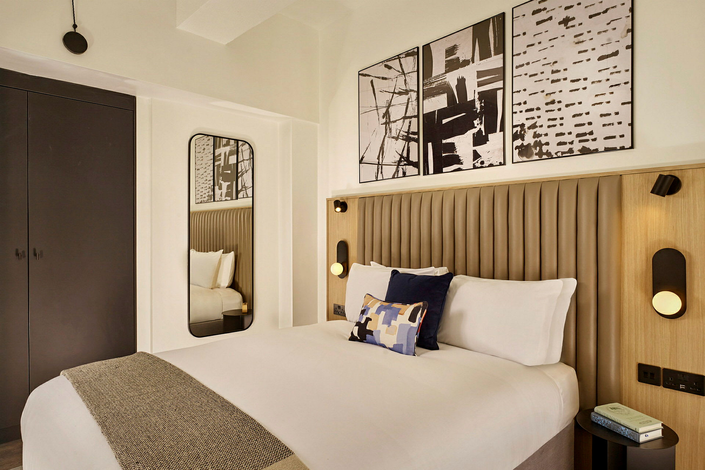 Ett modernt hotellrum med en säng klädd i vita sängkläder och dekorativa kuddar, en spegel på väggen och konstverk ovanför sängramen. (AI-genererad text)