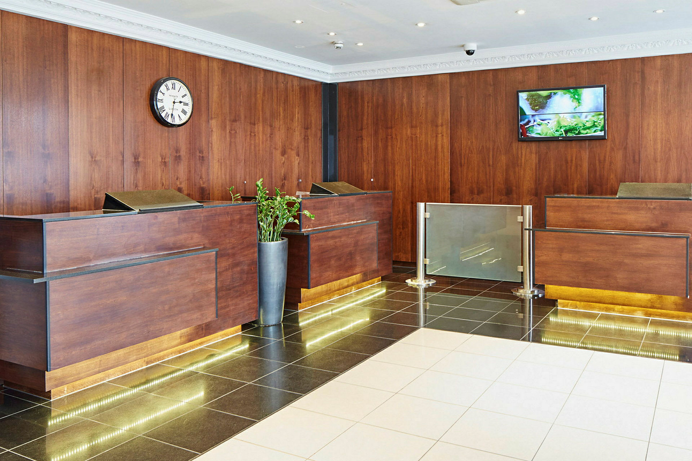 Reception med träpanel, skrivbord och klocka på väggen samt växt i kruka. (AI-genererad text)