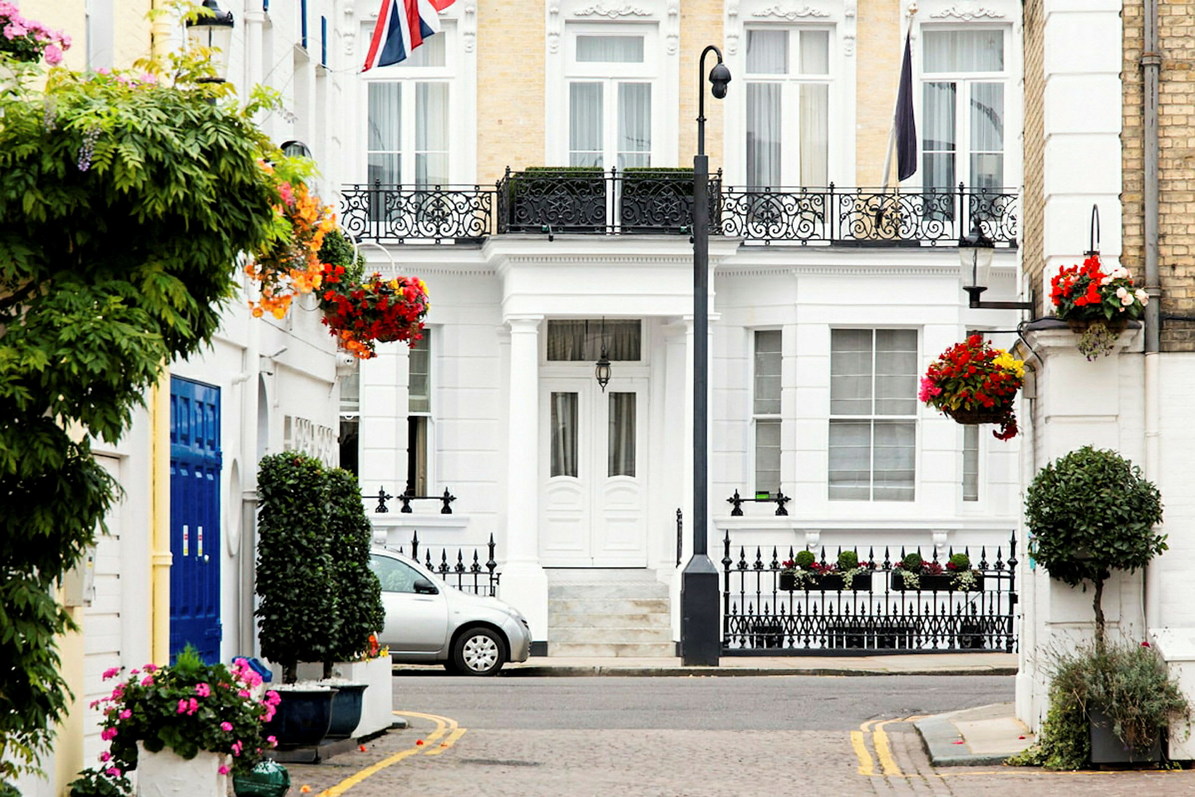 En vit byggnad med dekorativa järnräcken, blomsterarrangemang och en bil parkerad framför. Flaggan av Storbritannien syns. (AI-genererad text)