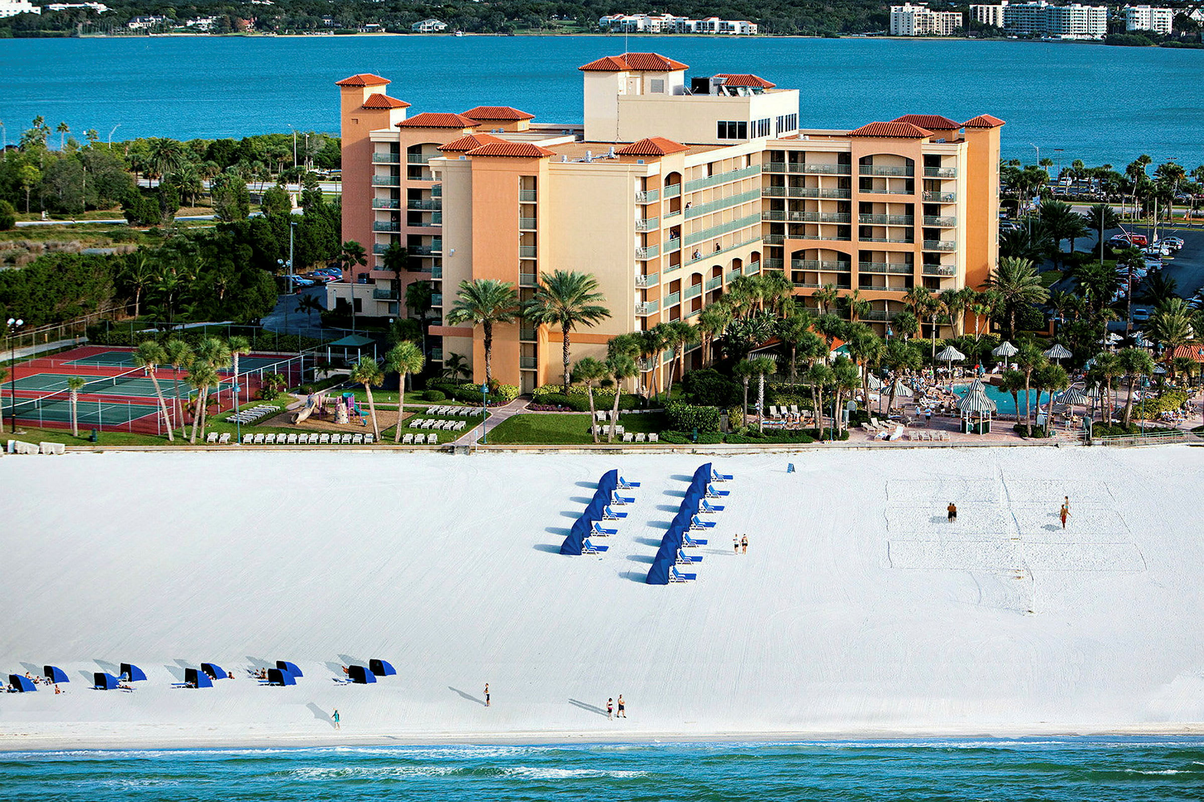 Ett hotellkomplex vid stranden, med blå parasoller och solstolar på den vita sanden, och en bakgrund av havet. (AI-genererad text)
