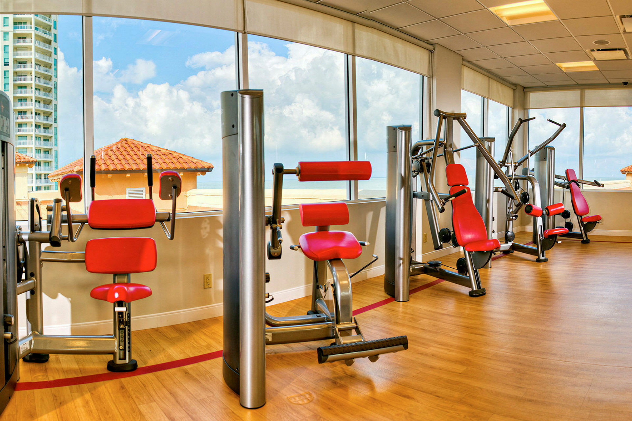 Ett gym med träningsmaskiner med röda säten och ryggstöd, placerade framför stora fönster som visar en vy av hustak och himmel. (AI-genererad text)
