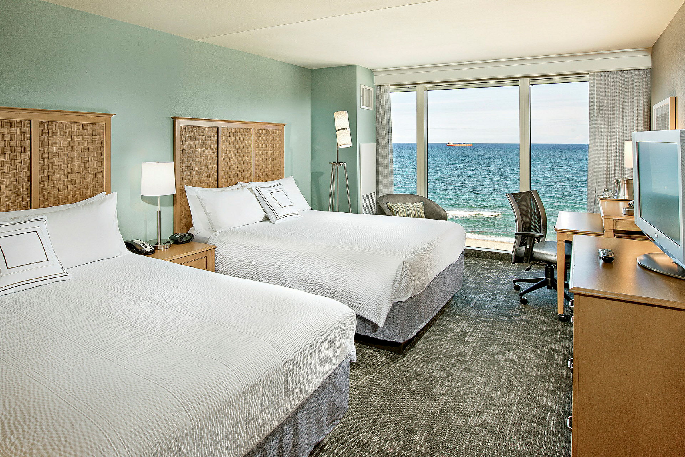 Hotellrum med två dubbelsängar, en TV på ett skrivbord och en stor glasvägg med utsikt över havet. (AI-genererad text)