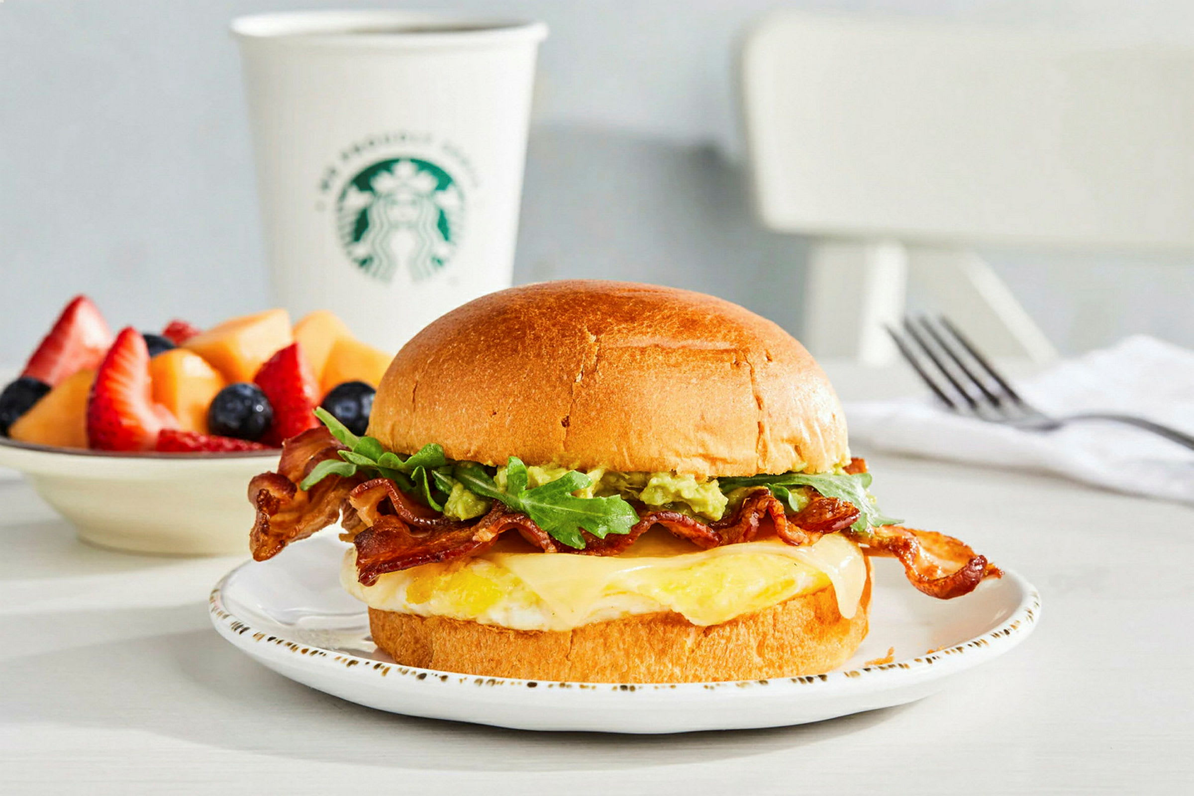 En tallrik med en macka innehållande bacon, sallad, ost och ett stekt ägg. I bakgrunden en kopp med märke och en skål med färsk frukt. (AI-genererad text)