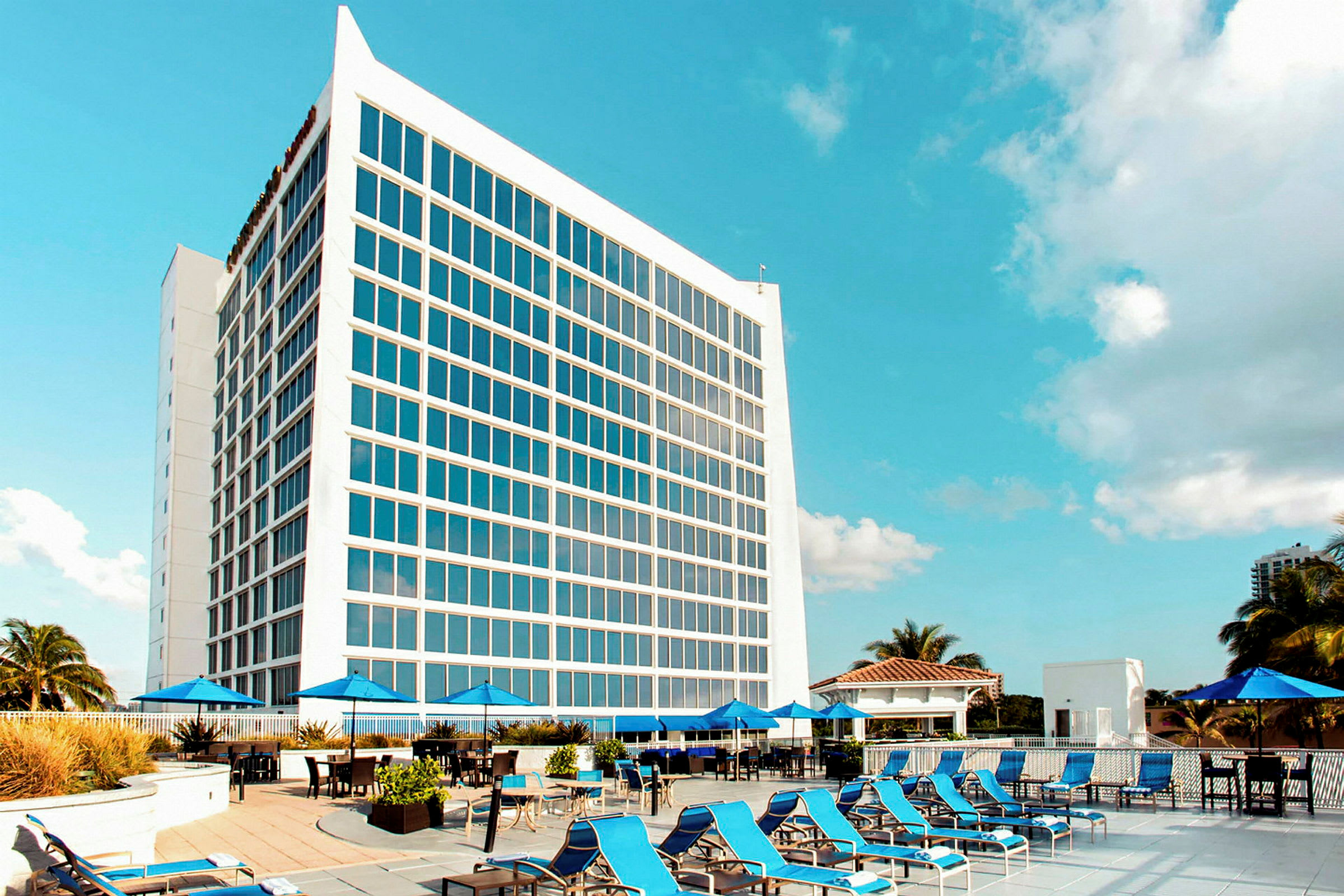 En modern hotellbyggnad med stora fönster bakom ett poolområde med solstolar och parasoller under en blå himmel. (AI-genererad text)