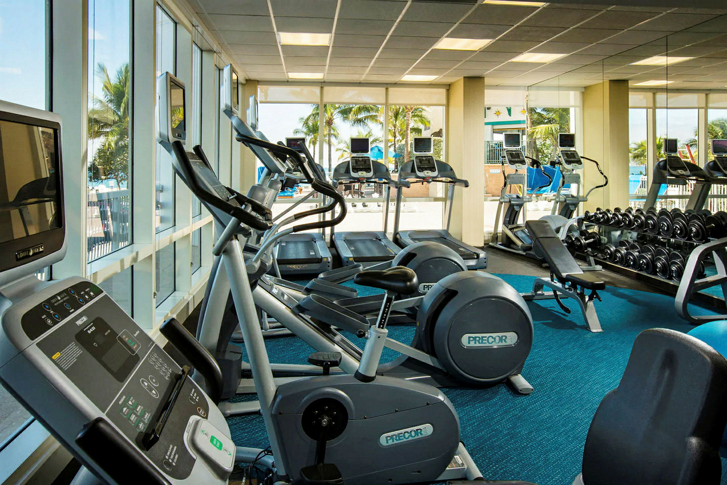 Ett gym med träningsmaskiner och fria vikter, med stora fönster som ger utsikt utomhus. (AI-genererad text)