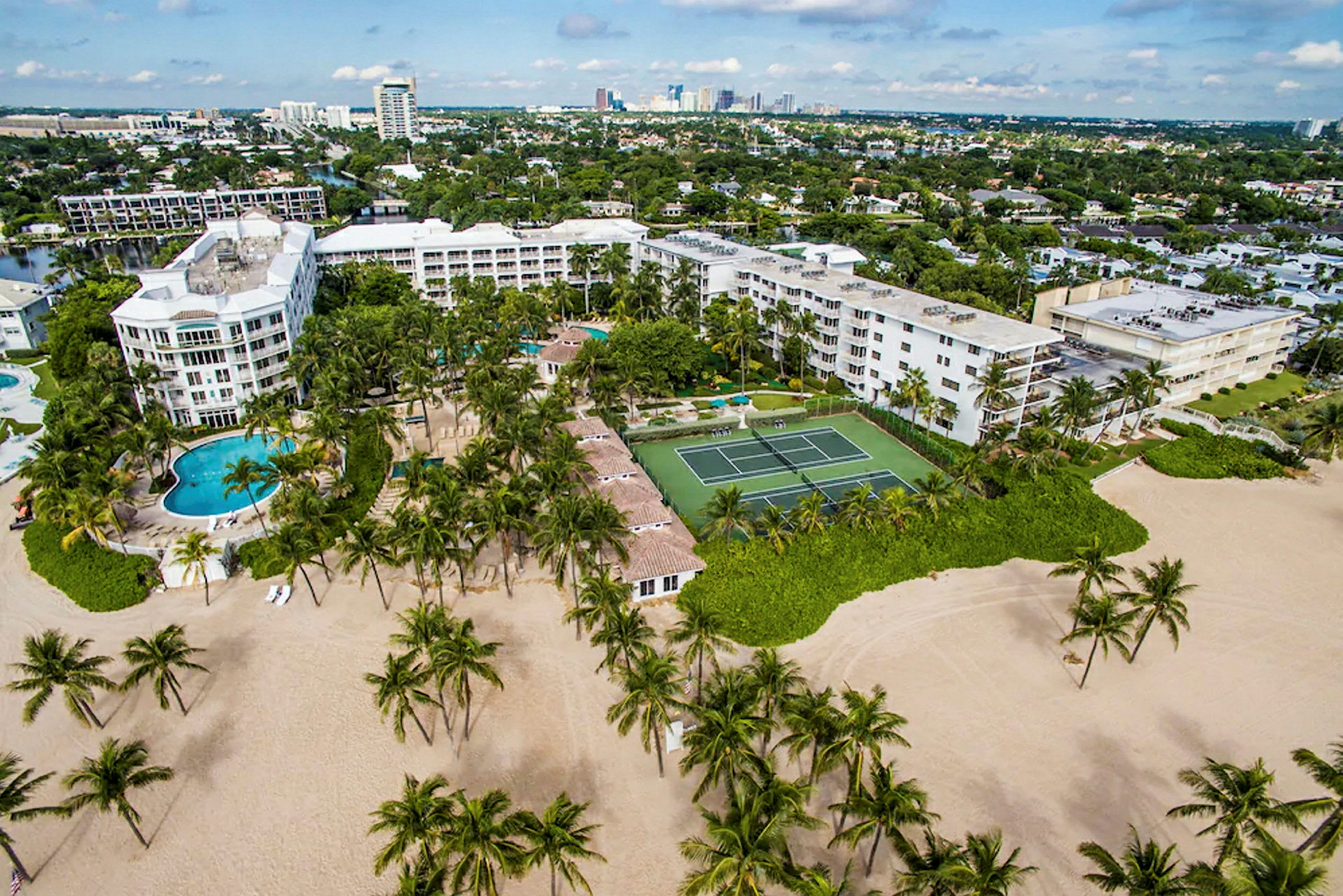 Flygbild över en semesteranläggning med pooler, tennisbana och omgivande byggnader, belägen nära en sandstrand med palmer. (AI-genererad text)
