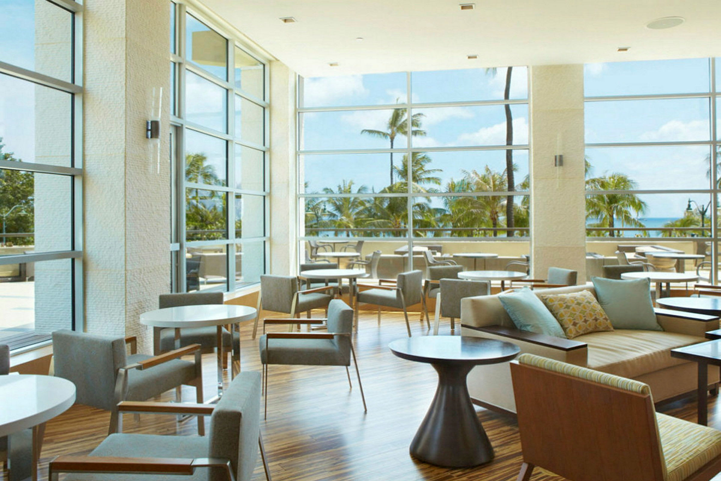 En hotellounge med stora glasfönster som erbjuder utsikt över palmer och havet. (AI-genererad text)