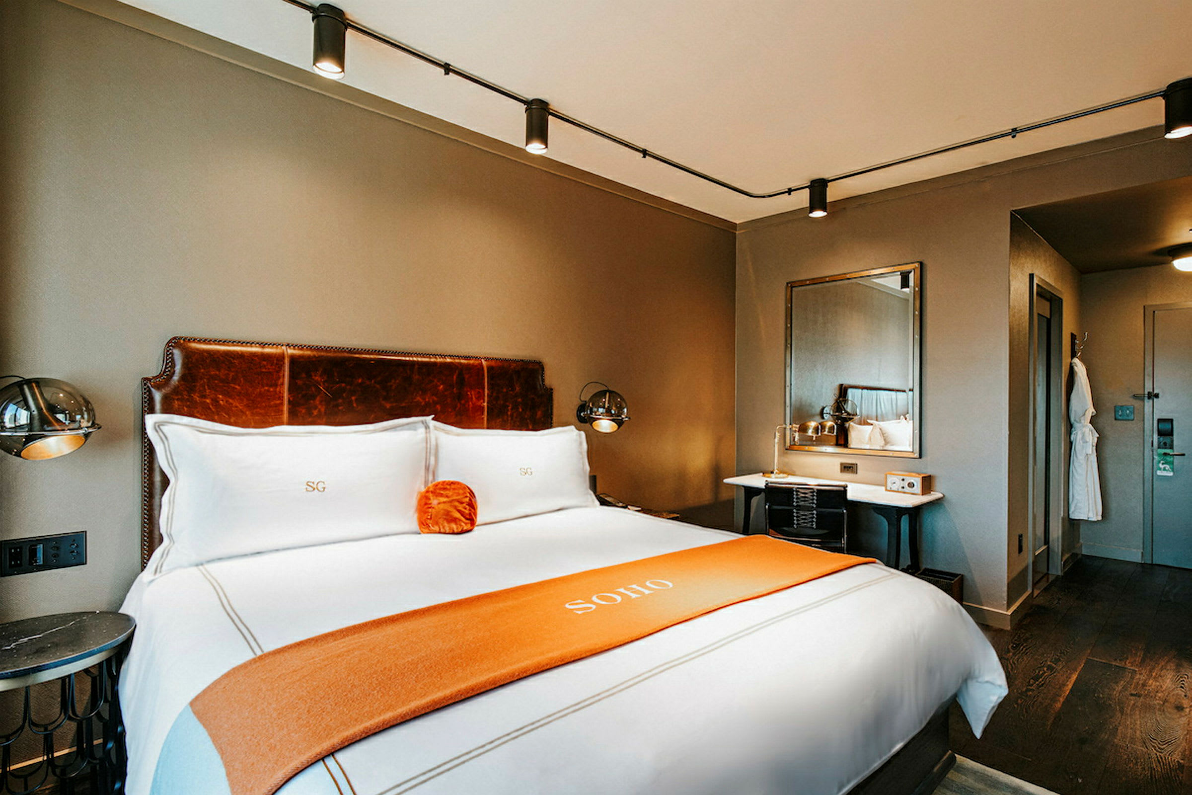 Ett hotellrum med en stor säng klädd i vita sängkläder och en orange filt. Ett skrivbord med spegel och lampa samt en badrock hänger på väggen. (AI-genererad text)