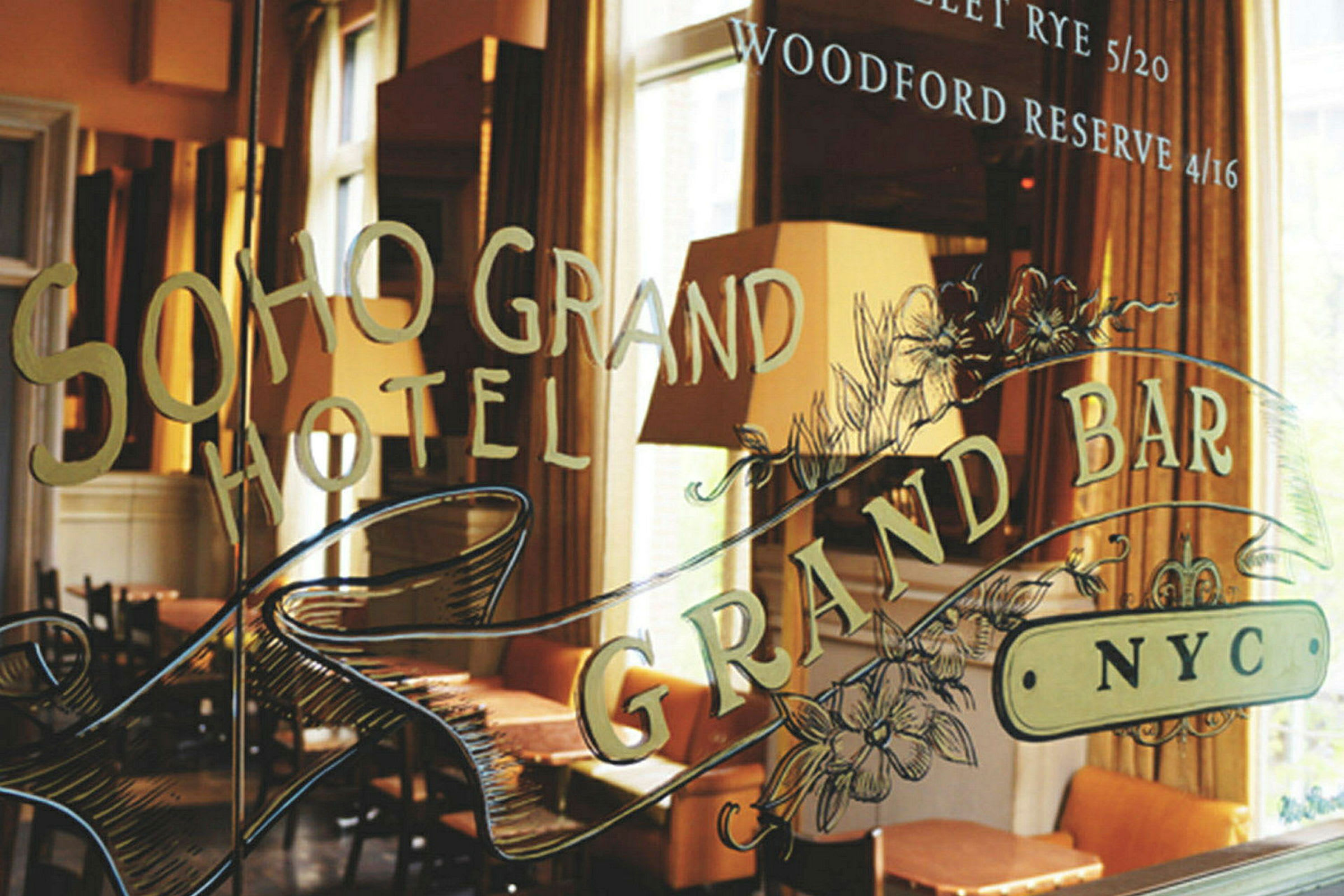 Ett fönster med texten 'Soho Grand Hotel' och 'Grand Bar NYC,' samt dekorationer. Bakgrunden visar möbler och lampor. (AI-genererad text)