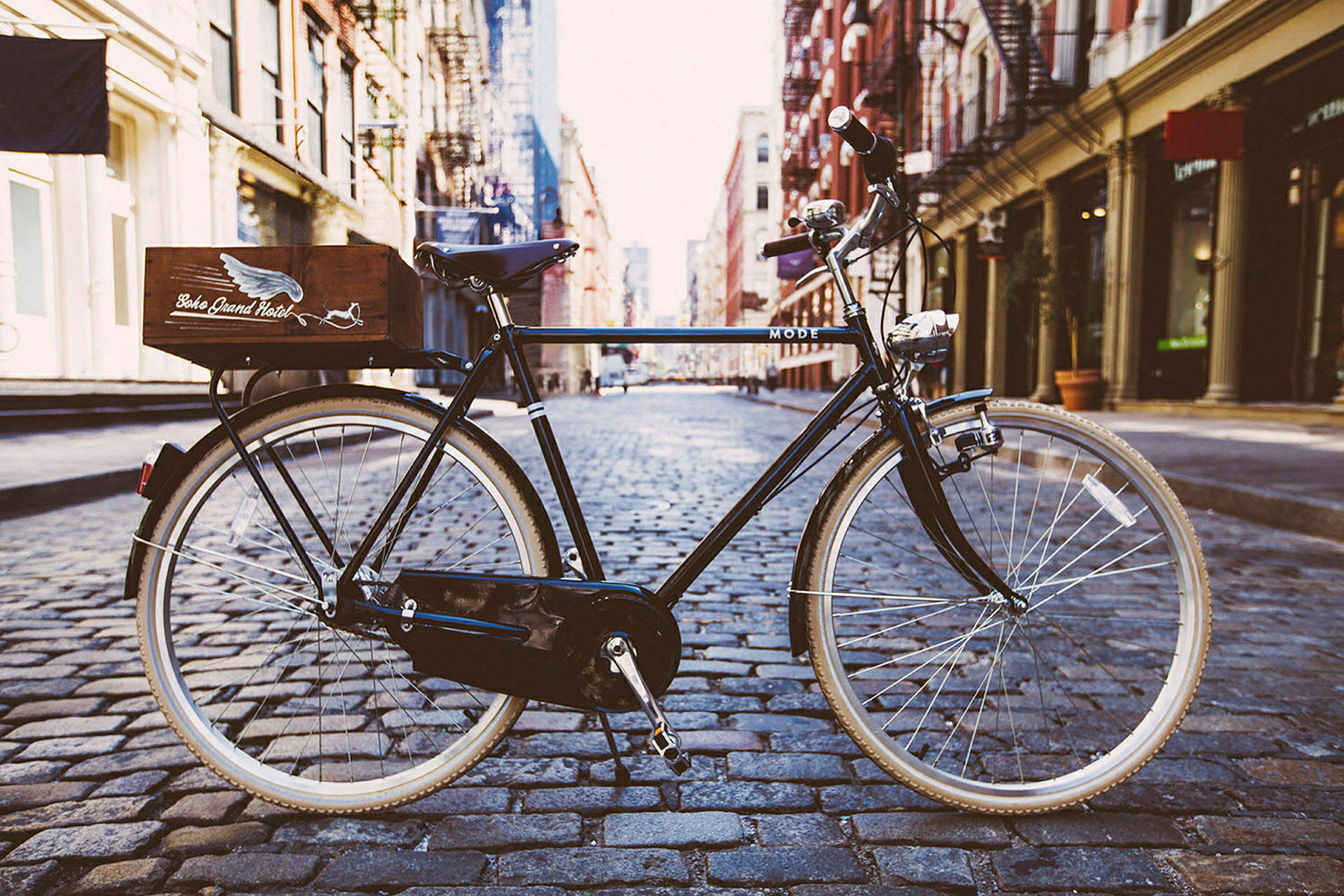 En svart cykel med en trälåda på bakre pakethållaren står på en stenlagd gata. I bakgrunden syns byggnader och trottoarer. (AI-genererad text)