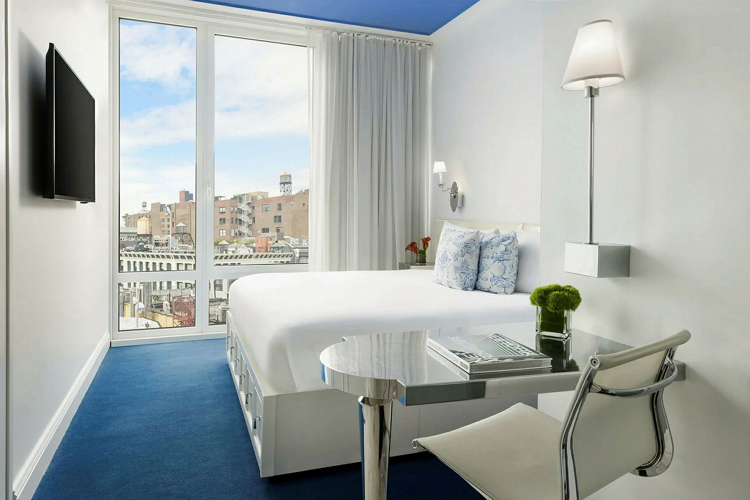 Ett ljust hotellrum med vit inredning och blå golv. En säng, ett skrivbord och en TV syns. (AI-genererad text)