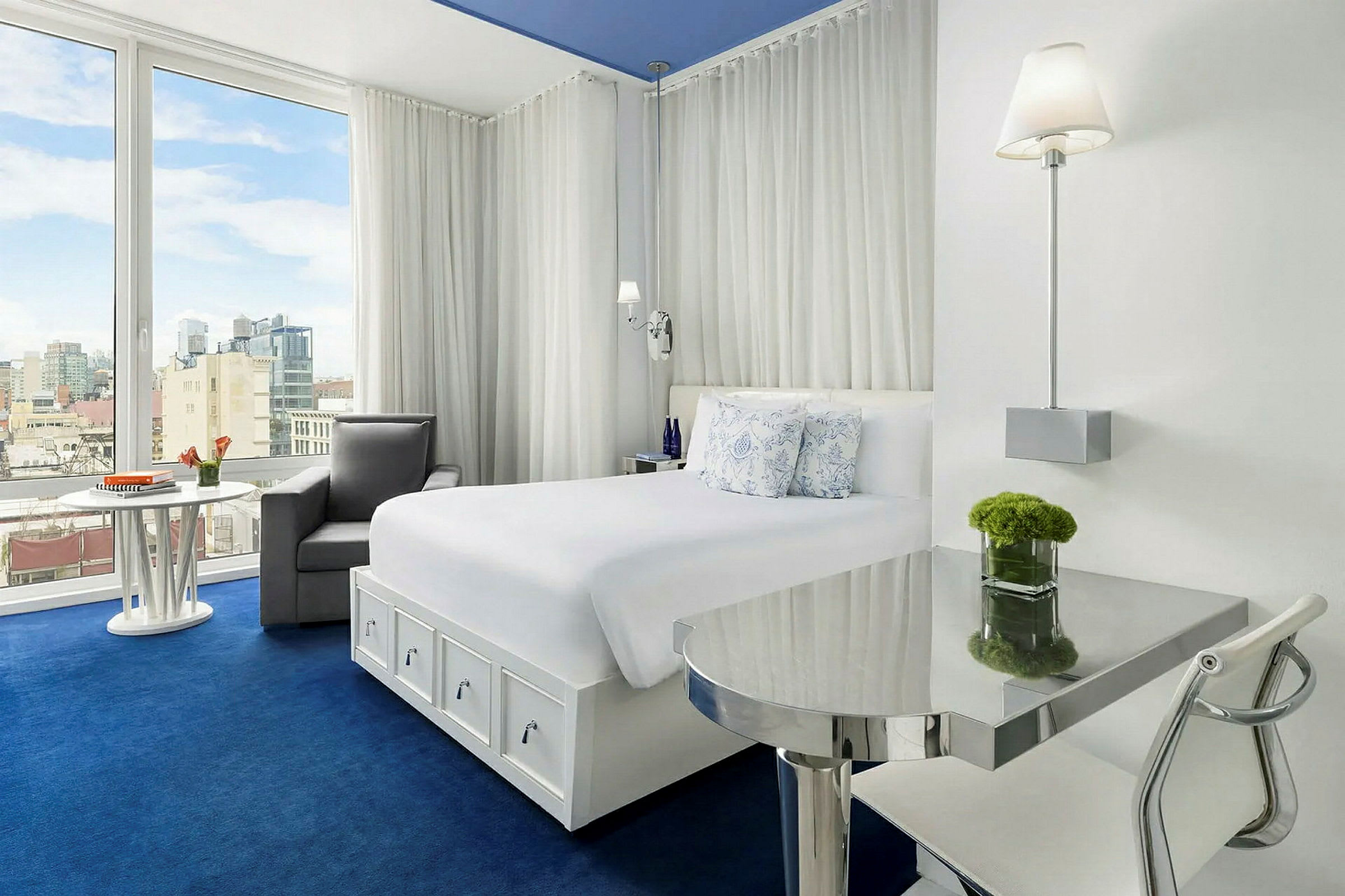 Ett ljust och modernt hotellrum med vit inredning och en stor säng. Rummet har ett fönster med utsikt över en stad. (AI-genererad text)