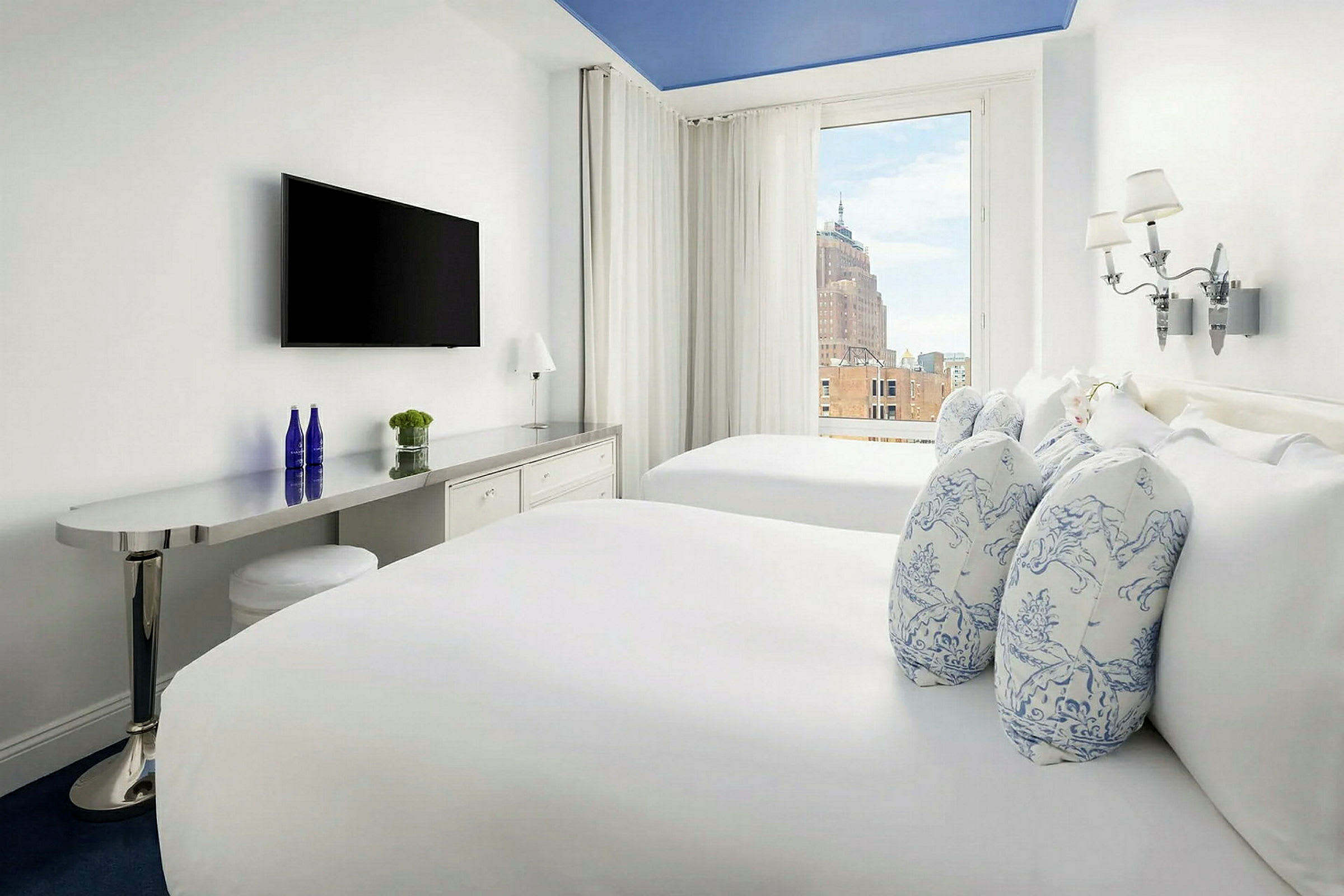 Hotellrum med två sängar med vita sängkläder och dekorativa kuddar, en väggmonterad TV, samt ett stort fönster med utsikt över byggnader. (AI-genererad text)