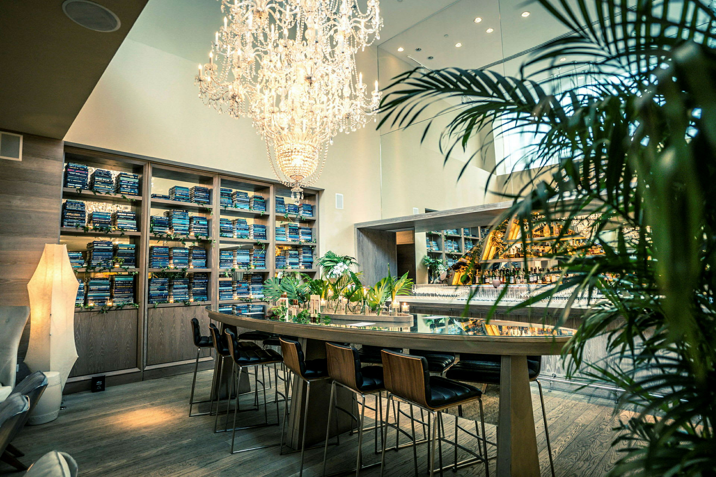 En elegant lokal med en kristallkrona ovanför ett långbord med stolar, ett barområde i bakgrunden och hyllor fyllda med böcker. (AI-genererad text)