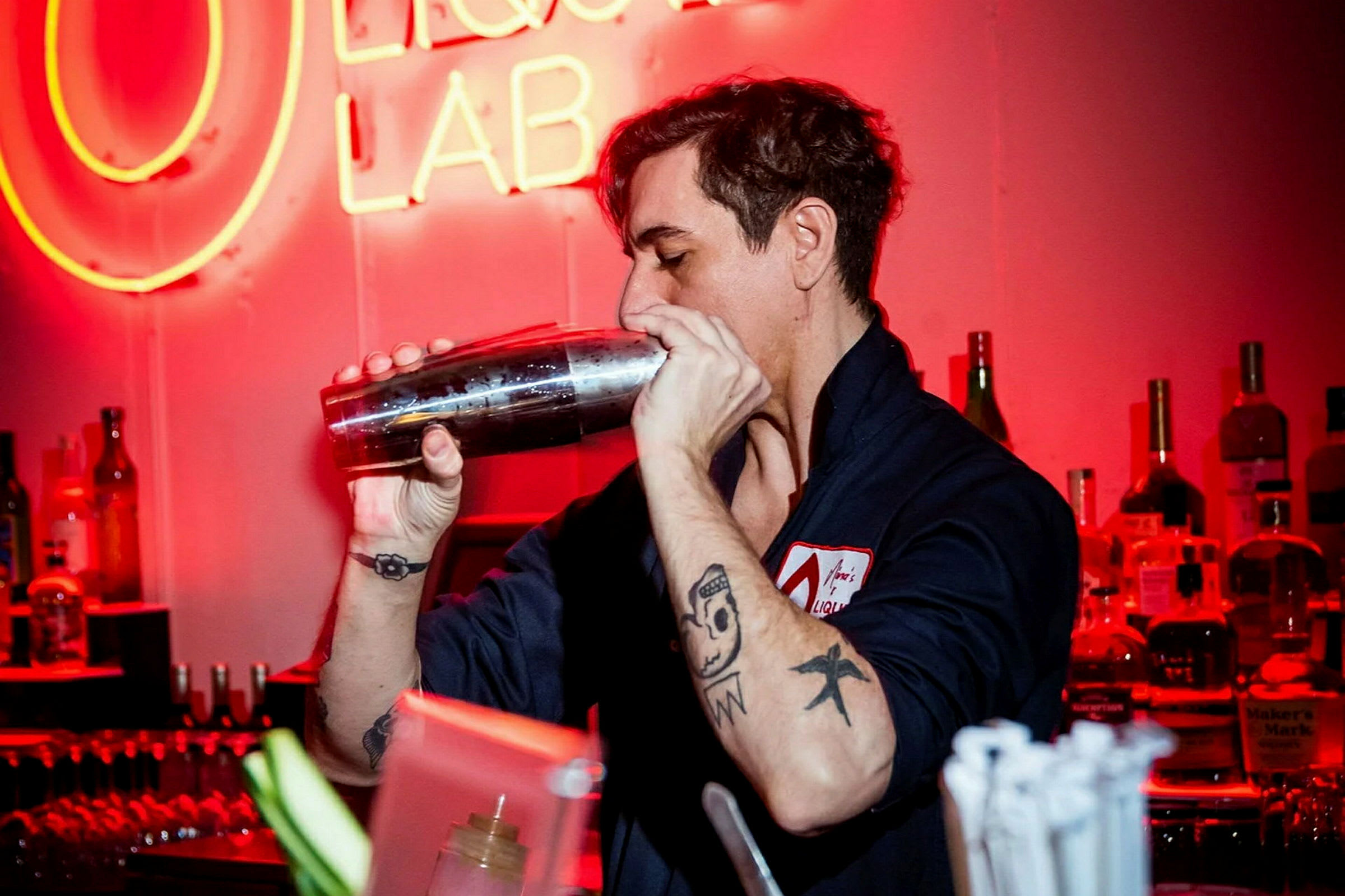 En person med tatueringar håller en cocktailshaker i en bar med röd belysning. (AI-genererad text)
