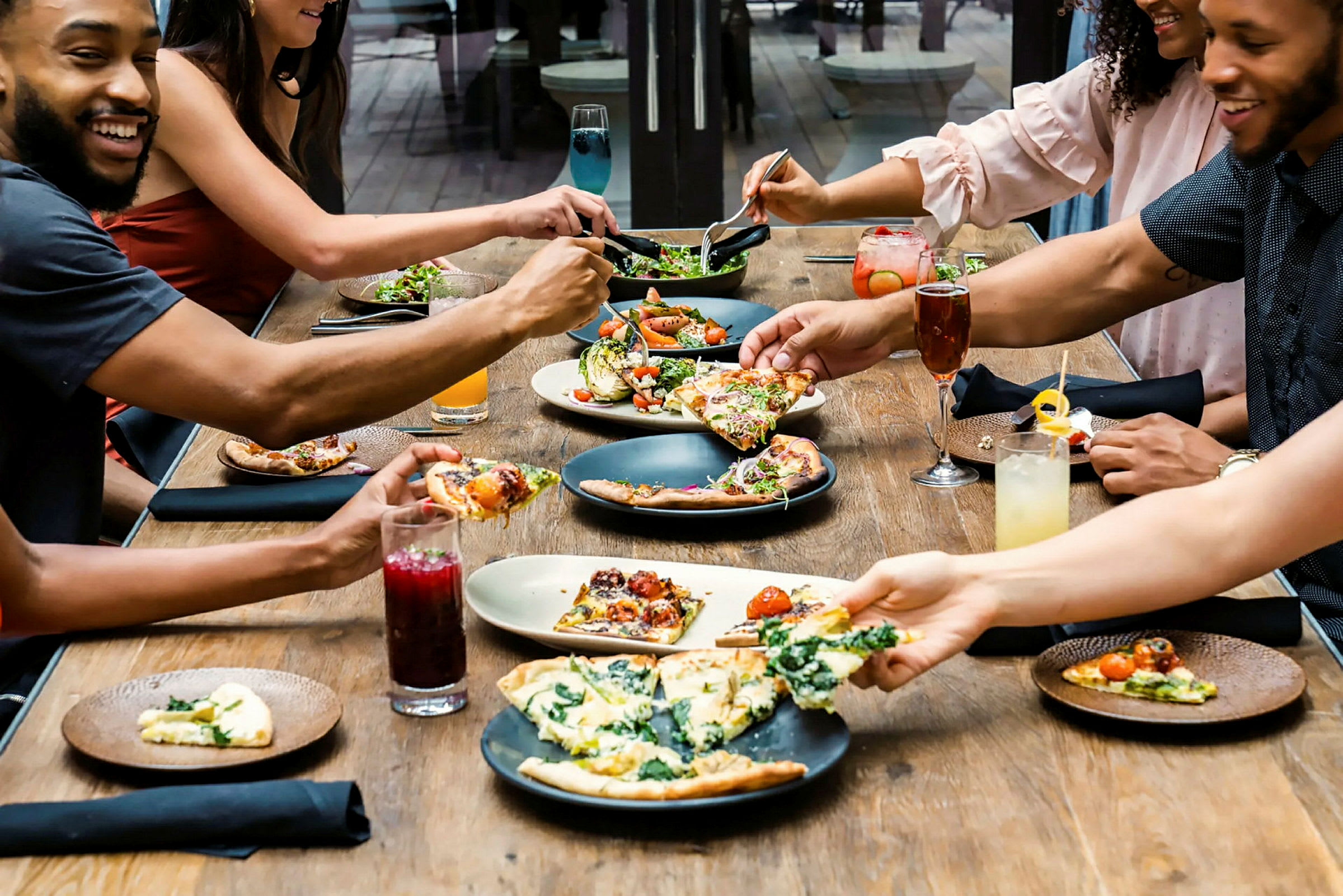 En grupp människor äter mat vid ett bord. Det serveras pizza och andra maträtter, tillsammans med olika drycker. (AI-genererad text)